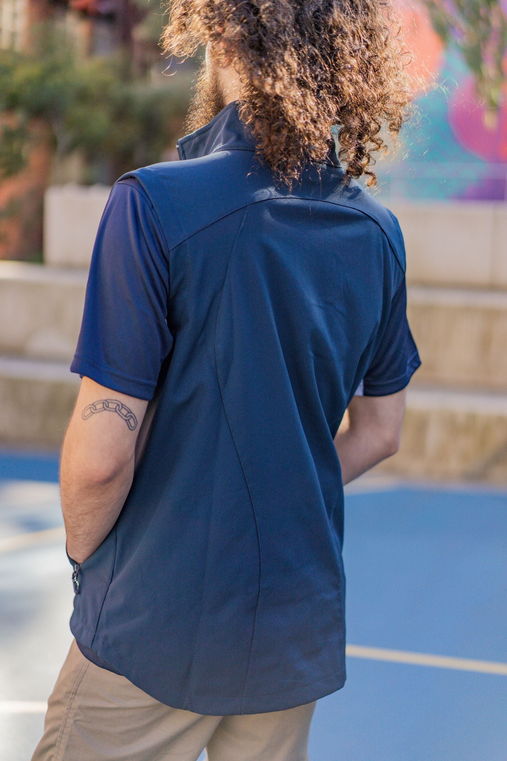 Rear view of male model wearing the navy Health, Physical Education & Sport Uniform – Vest over a matching polo shirt, standing outdoors on campus with arms by his sides; Available at the RMIT Store.