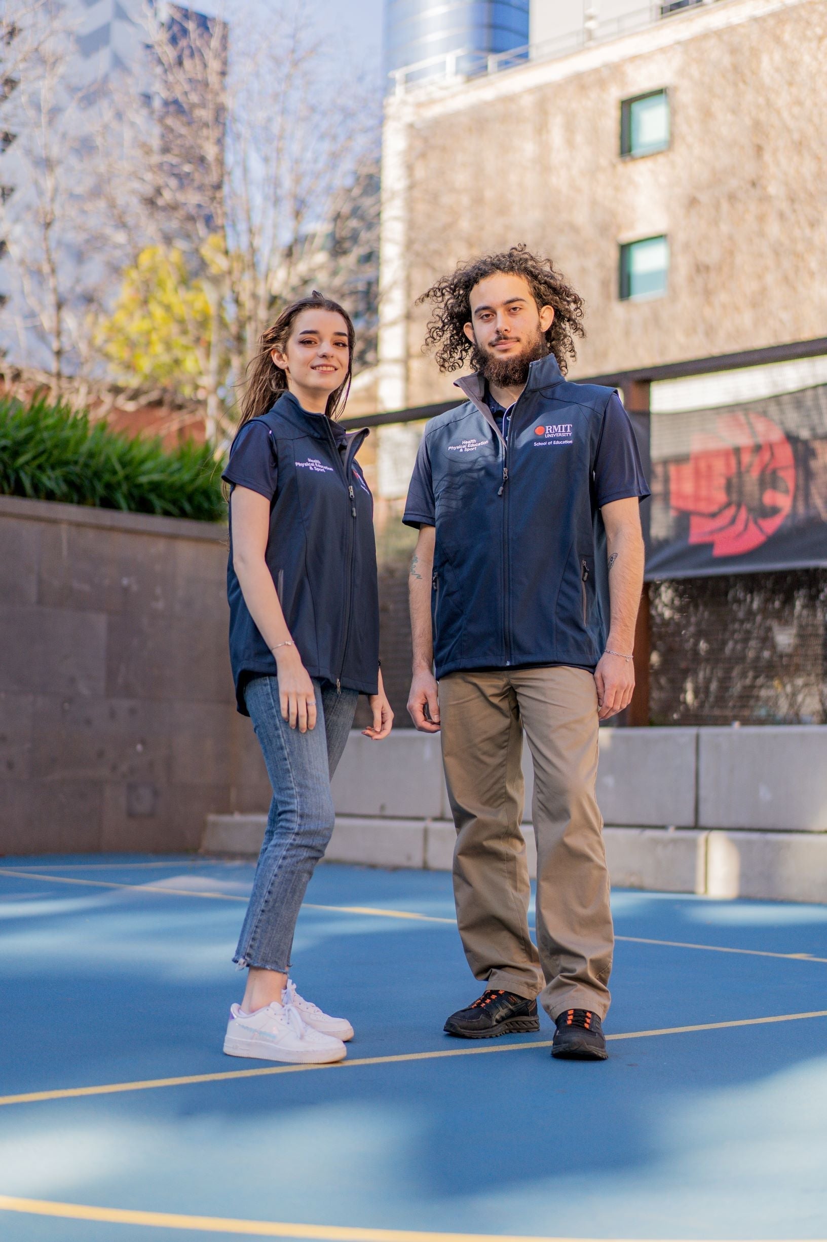 Front view of a male and female model both wearing the navy Health, Physical Education & Sport Uniform vest over matching navy Health, Physical Education & Sport Polo shirts from the RMIT Store, looking downwards outdoors on campus.