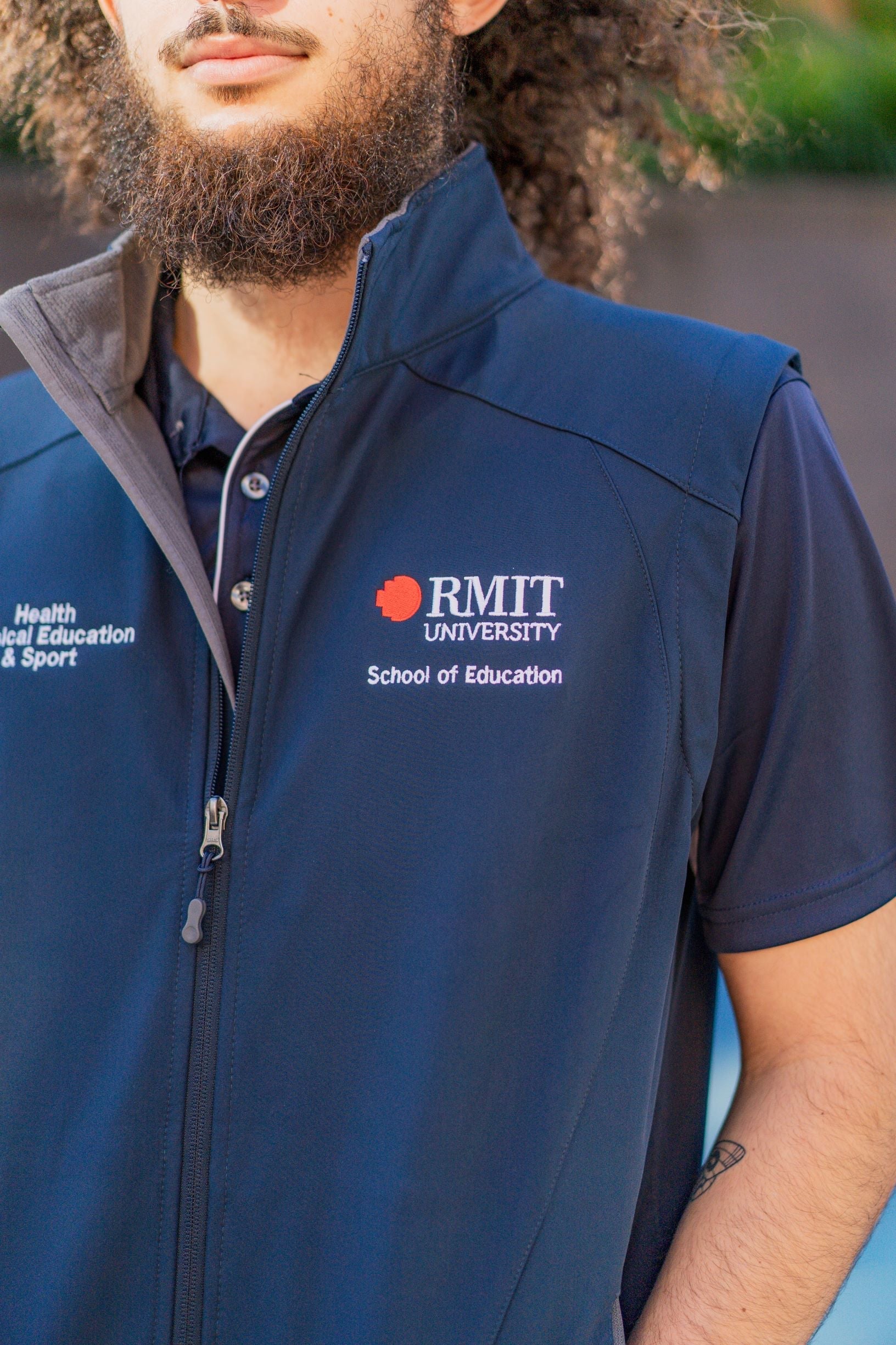 Close-up of the left chest of the navy Health, Physical Education & Sport Uniform – Vest worn by a male model, featuring white embroidered text reading "School of Education" beneath the RMIT University logo, captured in an outdoor campus setting. Available at the RMIT Store.