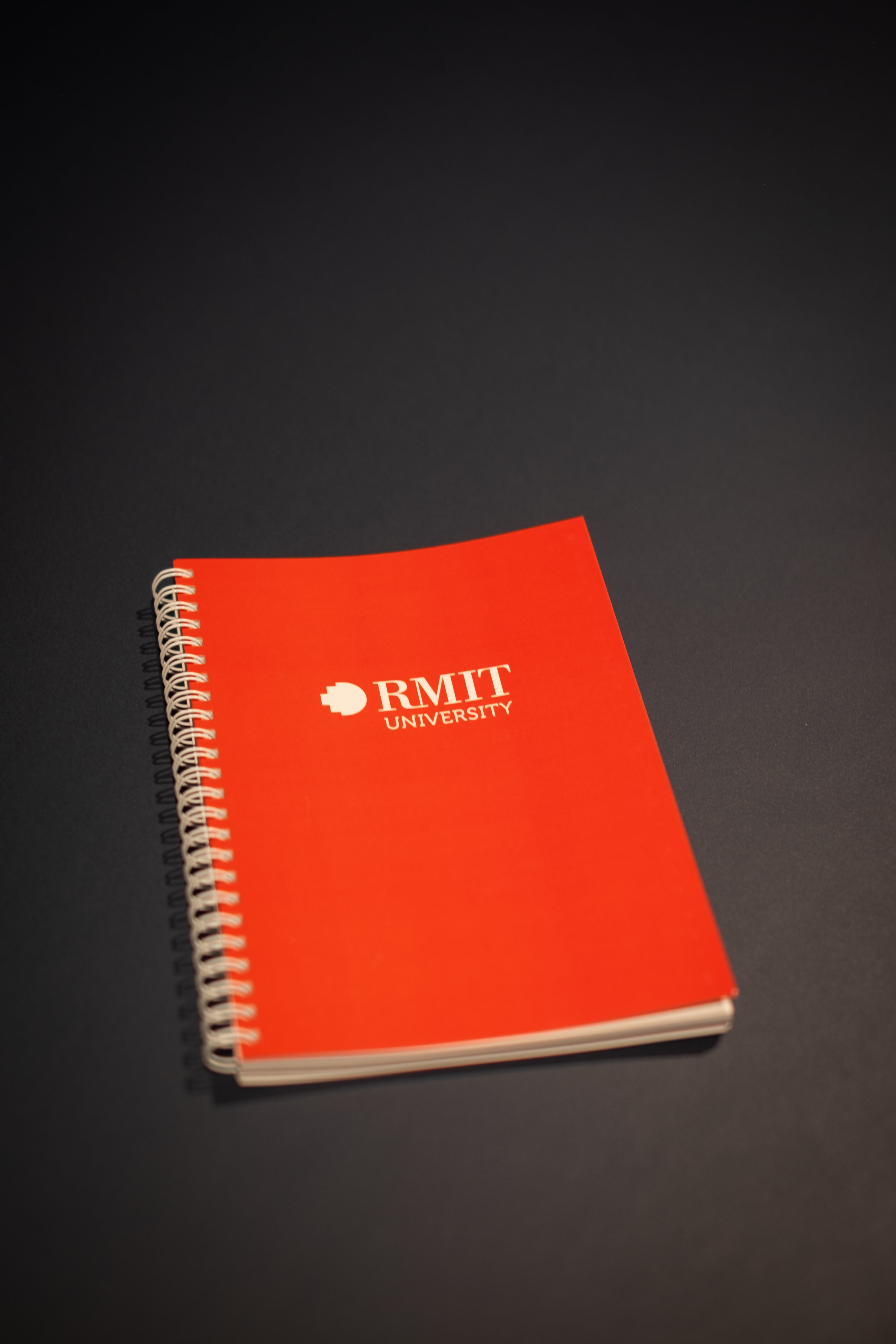 Interior Design and Decoration Kit including a red spiral-bound notebook displayed flat on a dark surface at RMIT Store, showing white printed RMIT University logo on the front cover, metal coil binding along the left edge, smooth matte cover finish, and visible stacked page edges.
