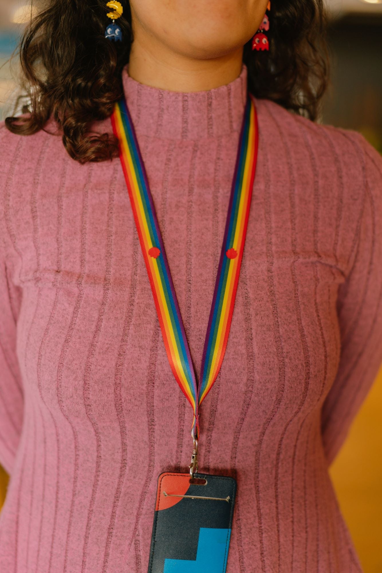 Front view of a female model wearing an Intersex-Inclusive Pride lanyard from the RMIT Store, featuring rainbow stripes with small red RMIT pixel logos. The lanyard is attached to a card holder. The model is shown from the chest down wearing a pink top.