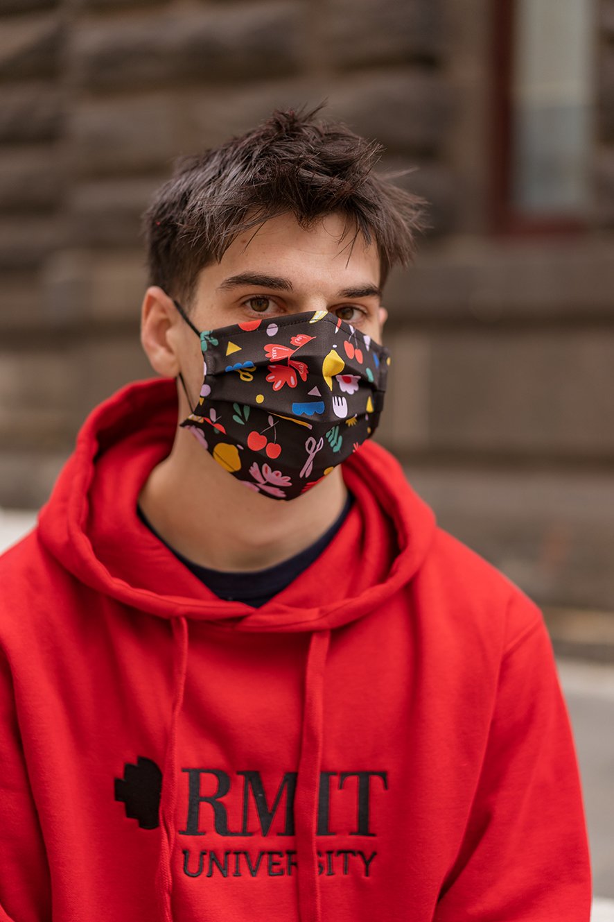 Male model wearing a red hoodie and an Isabella Manakis Designed Face Mask, showing a front view of the black fabric mask with subtle printed floral patterns, standing against a neutral background, from the RMIT Store.