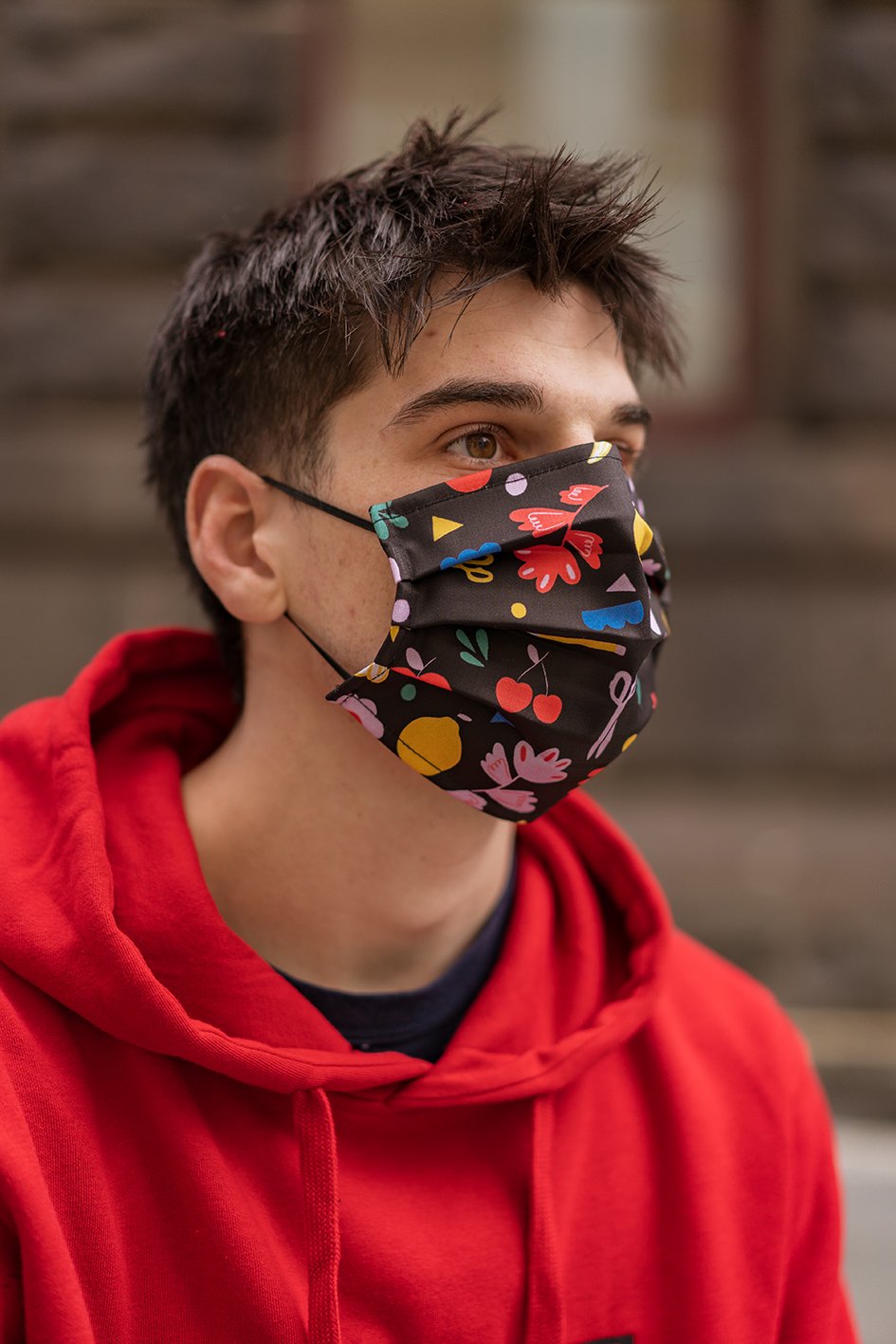 Male model in a red hoodie wearing an Isabella Manakis Designed Face Mask, showing a side view of the black fabric mask with faint floral prints, with the model looking slightly away from the camera, studio background, from the RMIT Store.
