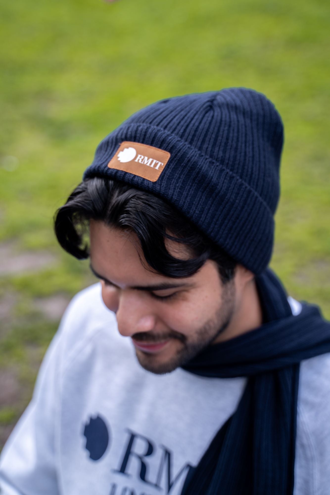 Close-up view of a male model wearing the navy RMIT knitted beanie with a brown PU patch embossed with the RMIT logo on the front. The model is looking down, showcasing the textured knit pattern and snug fit of the beanie. Available at the RMIT Store.