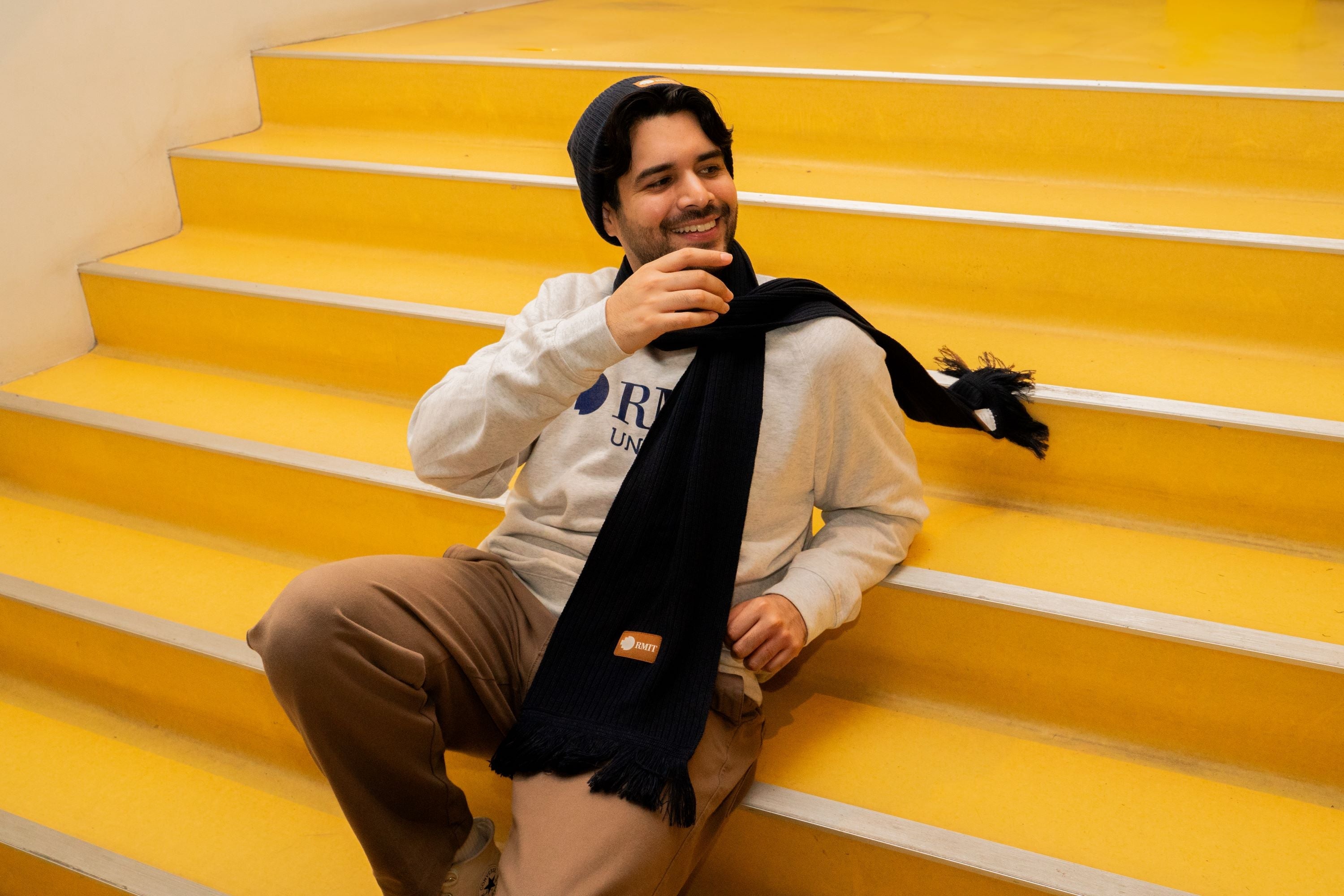Male model smiling and looking away from the camera, with one hand on his chin, wearing the navy RMIT knitted beanie and matching knitted scarf with textured patterns. He is dressed in a white crew neck sweater and seated outdoors on urban stairs. Available at the RMIT Store.