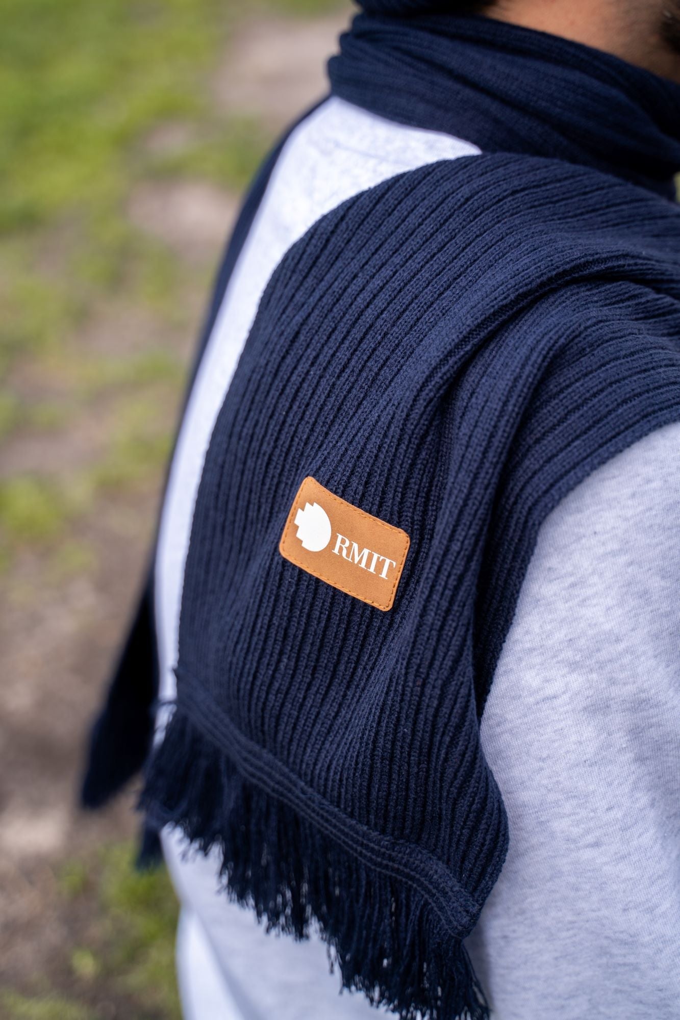 Close-up of the brown PU patch embossed with the RMIT logo, stitched onto the navy knitted scarf. The textured knit fabric of the scarf is clearly visible, showing tight, even stitching. Available at the RMIT Store.
