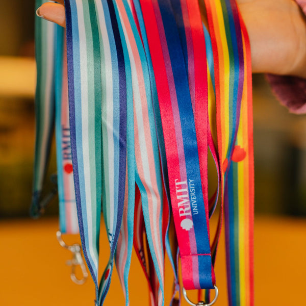 Close-up of a model’s hand holding a bundle of RMIT Store’s LGBTIQA+ Pride lanyards, including bisexual, transgender, pansexual, lesbian, intersex-inclusive, and trans-inclusive gay men’s designs. Each lanyard features distinct horizontal stripes and silver metal clips with subtle engraved RMIT logos near the attachment points