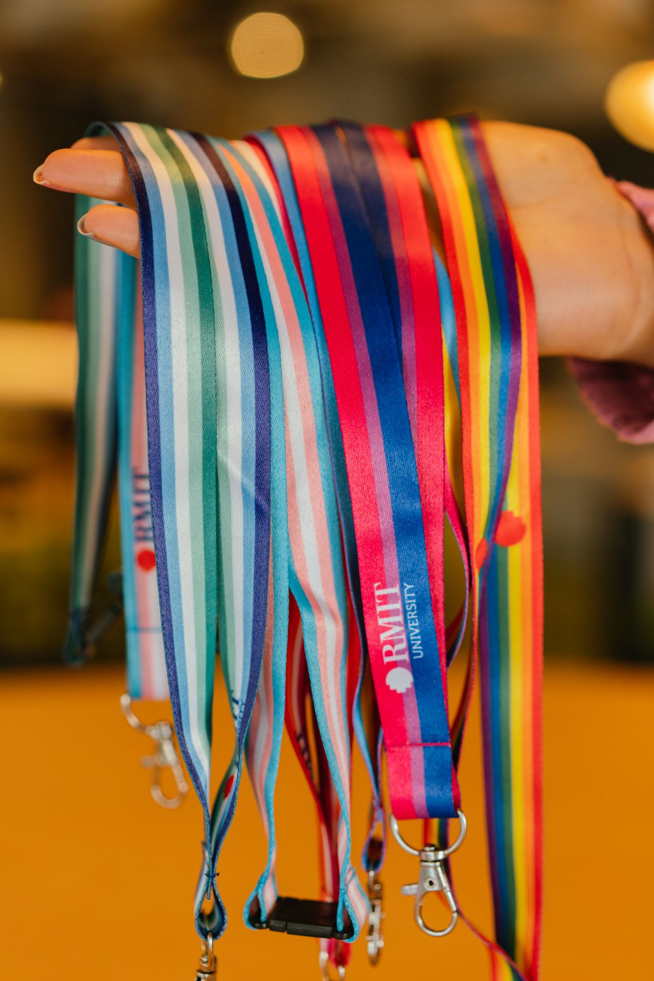 Close-up of a model’s hand holding a bundle of RMIT Store’s LGBTIQA+ Pride lanyards, including bisexual, transgender, pansexual, lesbian, intersex-inclusive, and trans-inclusive gay men’s designs. Each lanyard features distinct horizontal stripes and silver metal clips with subtle engraved RMIT logos near the attachment points