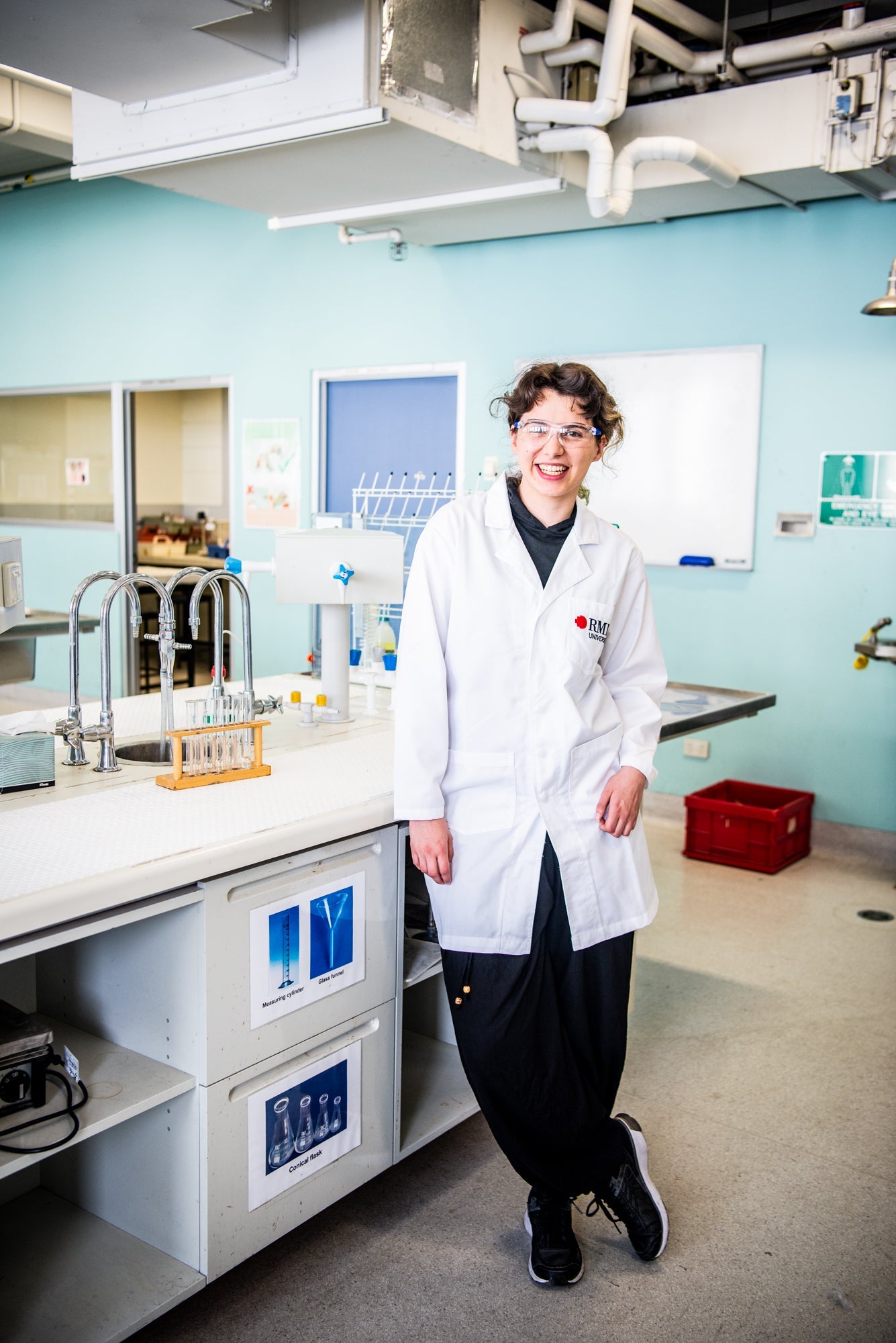 RMIT Store white lab coat worn by a model standing in a laboratory setting, showcasing the front design with pockets and the RMIT University logo.