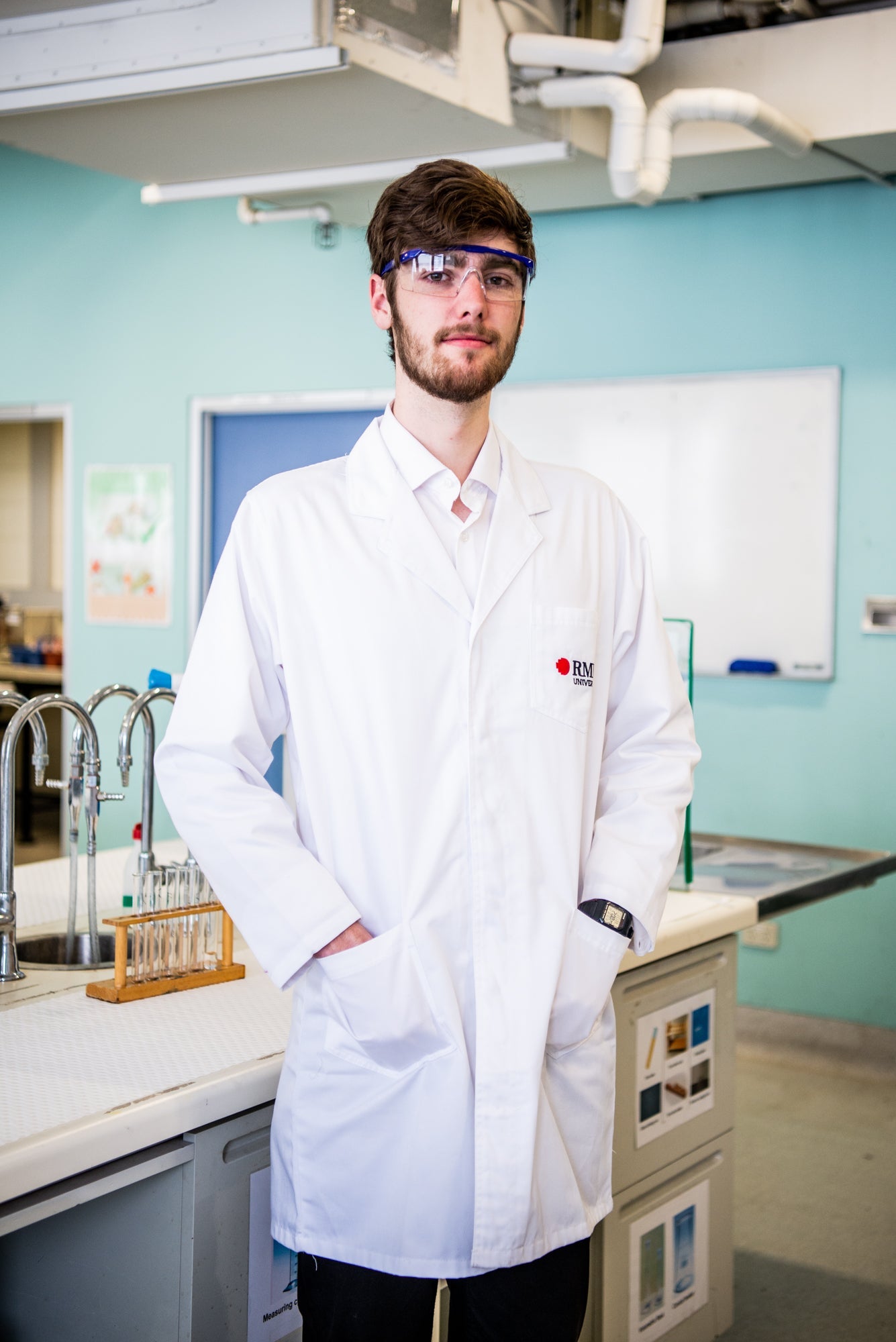 A front view of a model in a white lab coat from the RMIT Store, standing in a laboratory. The coat features the RMIT logo on the chest, with pockets visible. The lab is equipped with benches and test tubes under bright lighting.