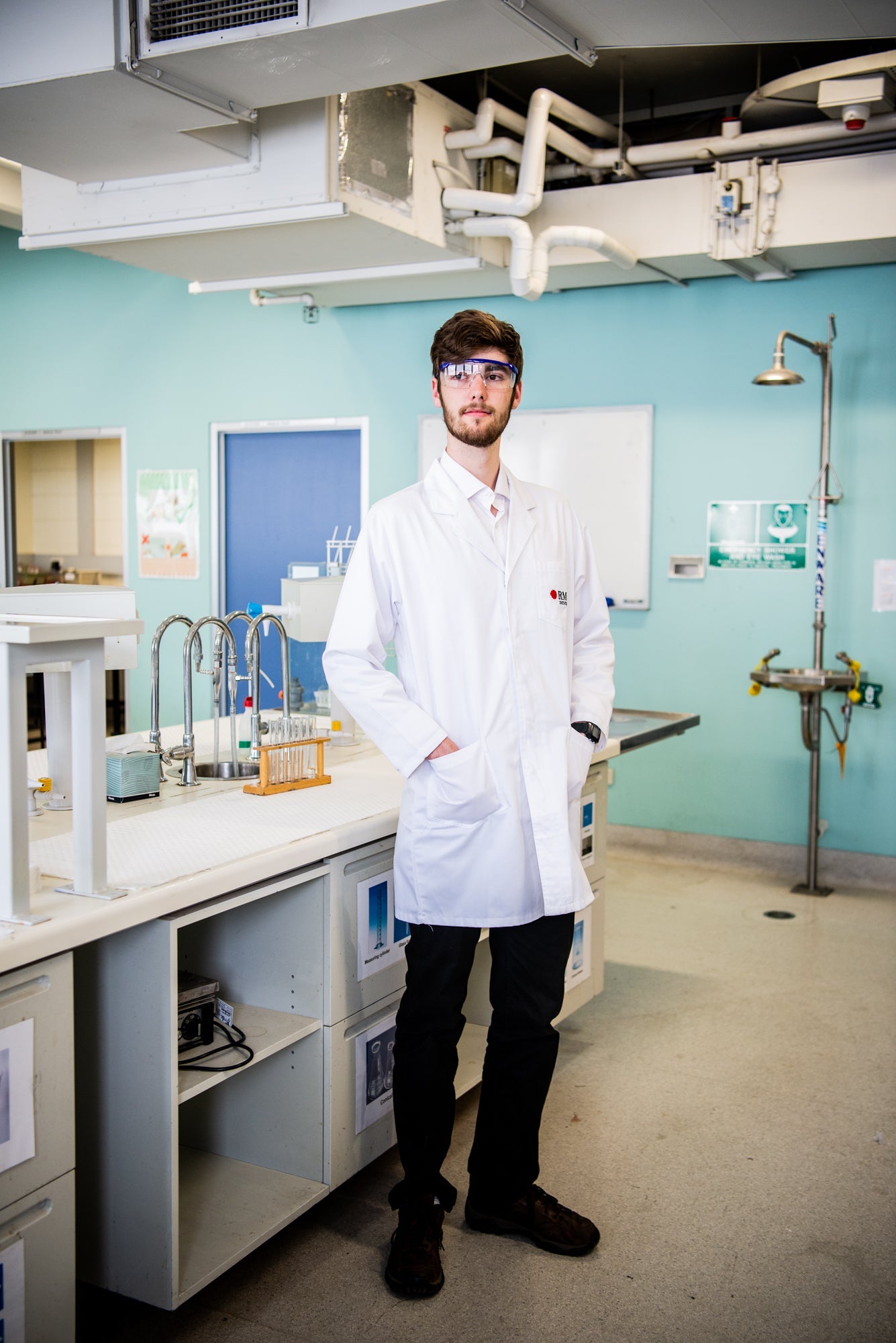 A front view of a model in a white lab coat from the RMIT Store, standing in a laboratory. The coat features the RMIT logo on the chest, with pockets visible. The lab is equipped with benches and test tubes under bright lighting.