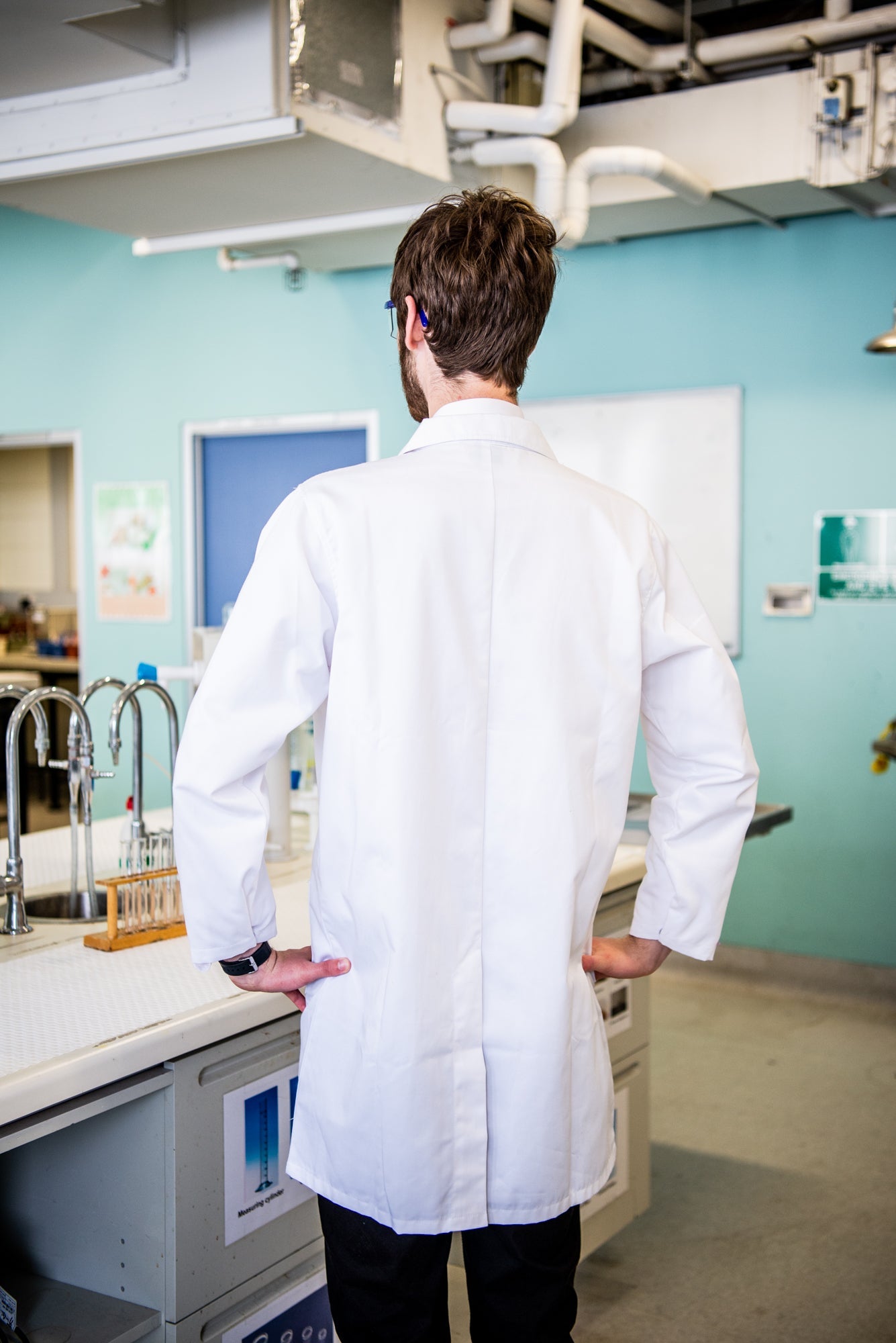 A model wearing a white lab coat from the RMIT Store standing in a laboratory. The back view highlights the coat's length and form, set within a science lab featuring equipment and bright blue walls.