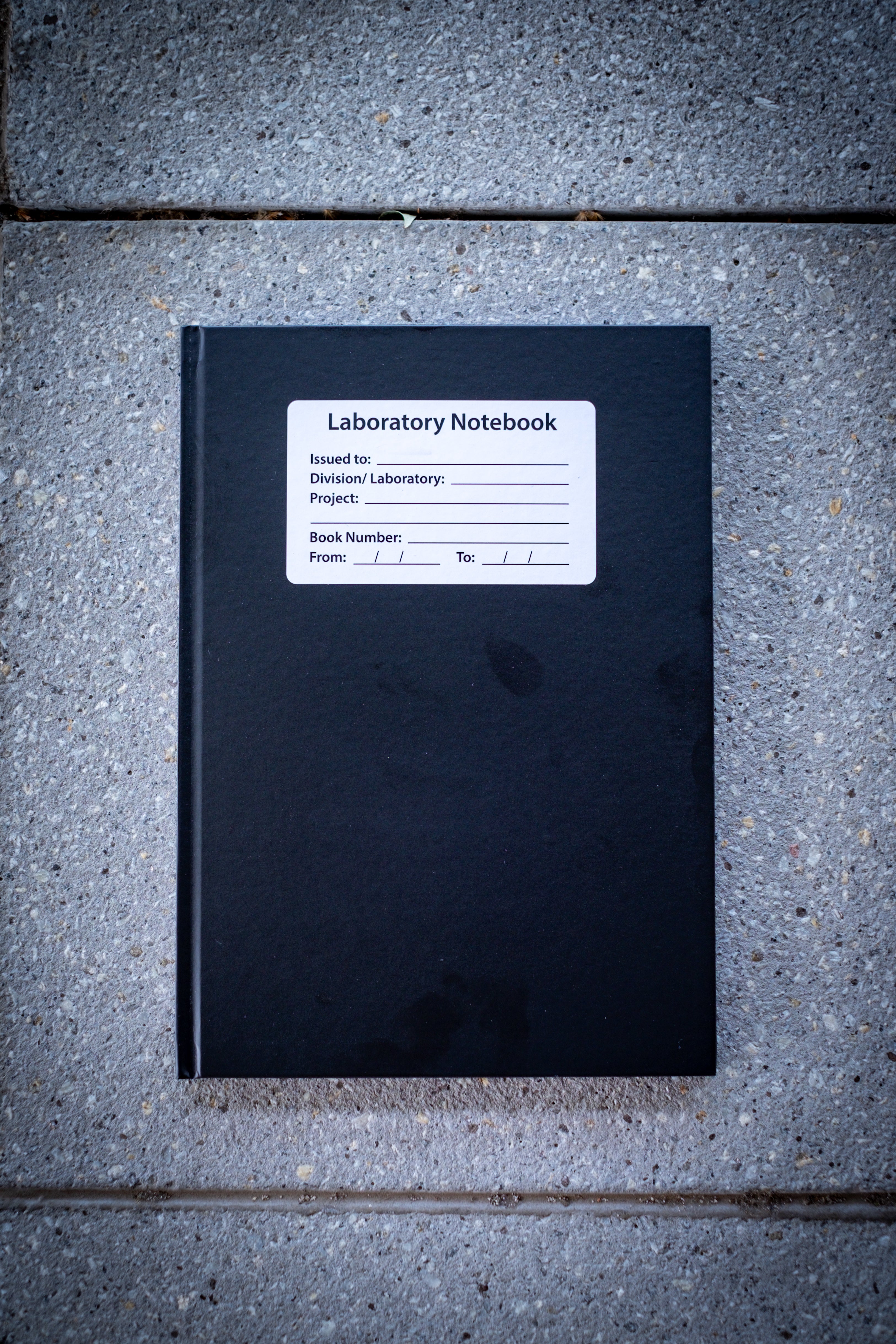 Laboratory Notebook in black variant shown closed on a light surface, featuring a matte black cover with rounded corners and a blank white rectangular label centred on the front — a durable and practical academic notebook from RMIT Store.