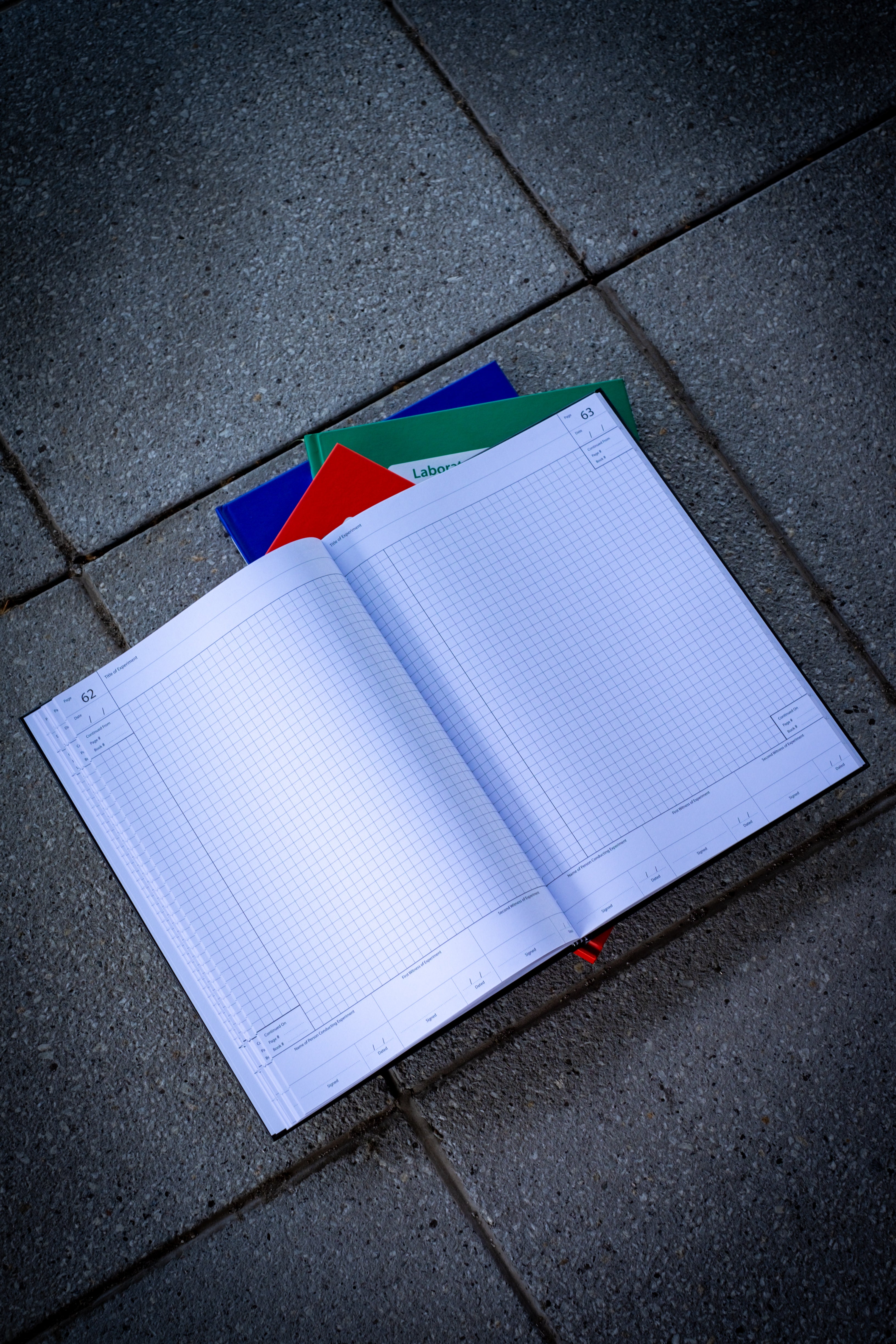 Opened Laboratory Notebook in black variant displayed on a light surface, showing its grid pages. Beneath it are three closed notebooks — red, blue, and green variants,