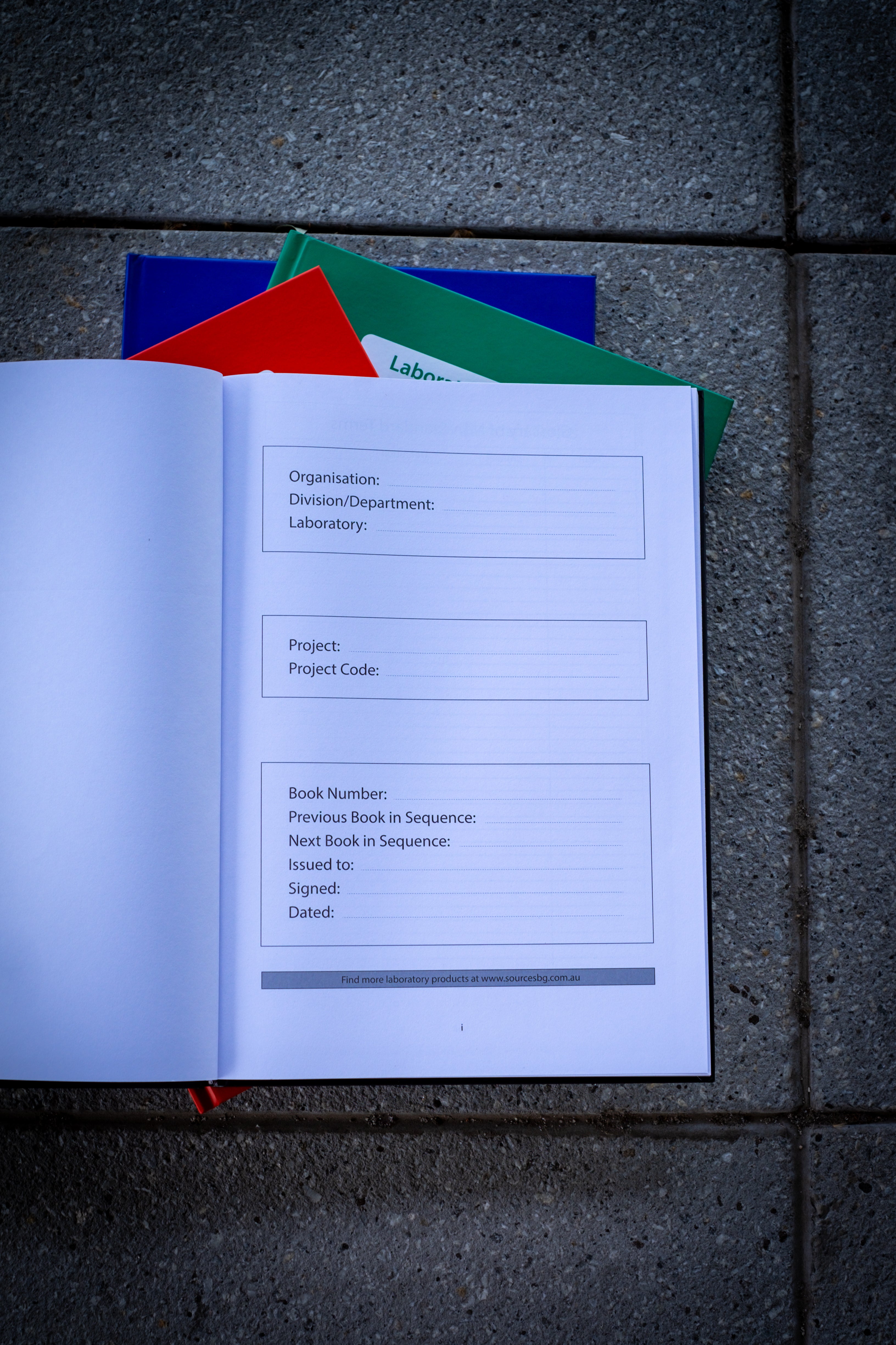 A close-up of a black Laboratory Notebook open on a desk, displaying pages with squares for customisation. The notebook is placed flat, and you can see the inside pages clearly. Beneath it, three additional closed notebooks (red, blue, and green) are stacked together. The focus is on the grid layout inside the black notebook, highlighting the notebook's structure and quality. Available at RMIT Store.