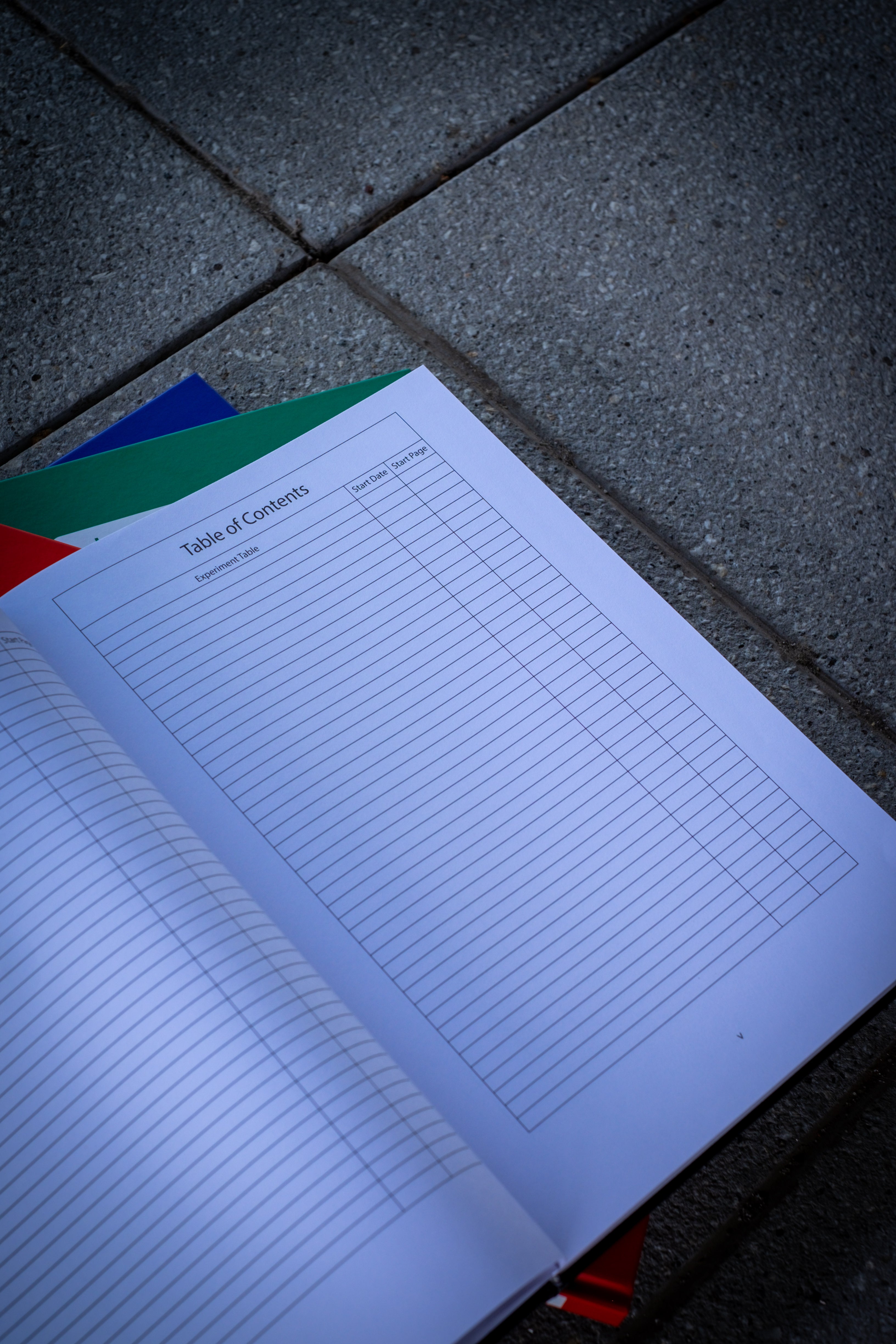 An opened black Laboratory Notebook on top, displaying the page titled "Table of Contents." The page features neatly organised text with grid lines visible in the background. Beneath the black notebook, three closed notebooks in red, blue, and green are stacked, with their spines aligned. The set is arranged on a black surface, showcasing the professional and organised design of the collection. Available at RMIT Store.