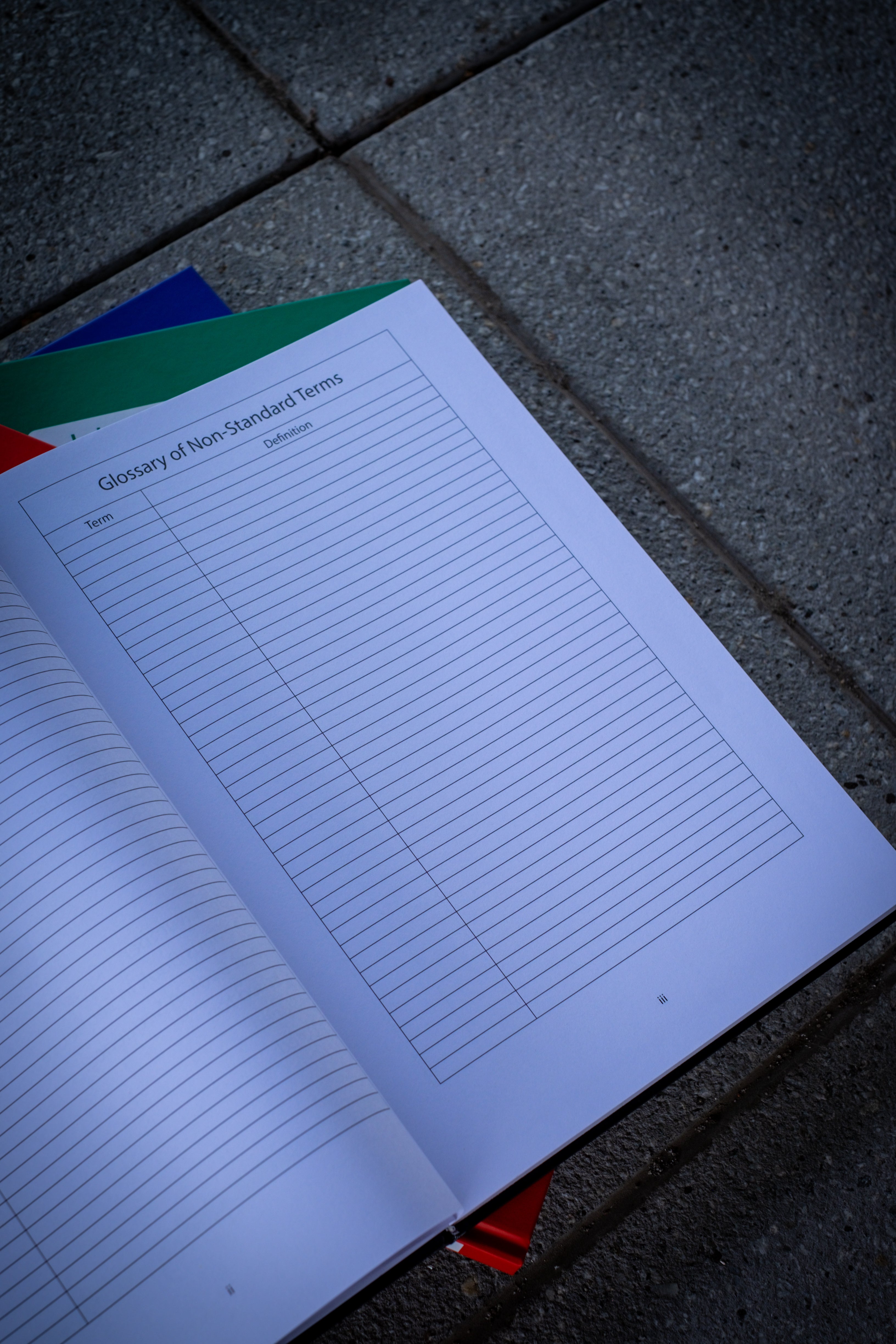 An opened black Laboratory Notebook on top, displaying the page titled "Glossary of Non-Standard Items." The page features neatly organised text with grid lines visible in the background. Beneath the black notebook, three closed notebooks in red, blue, and green are stacked. The set is arranged on a stone surface, showcasing the professional and organised design of the collection. Available at RMIT Store.