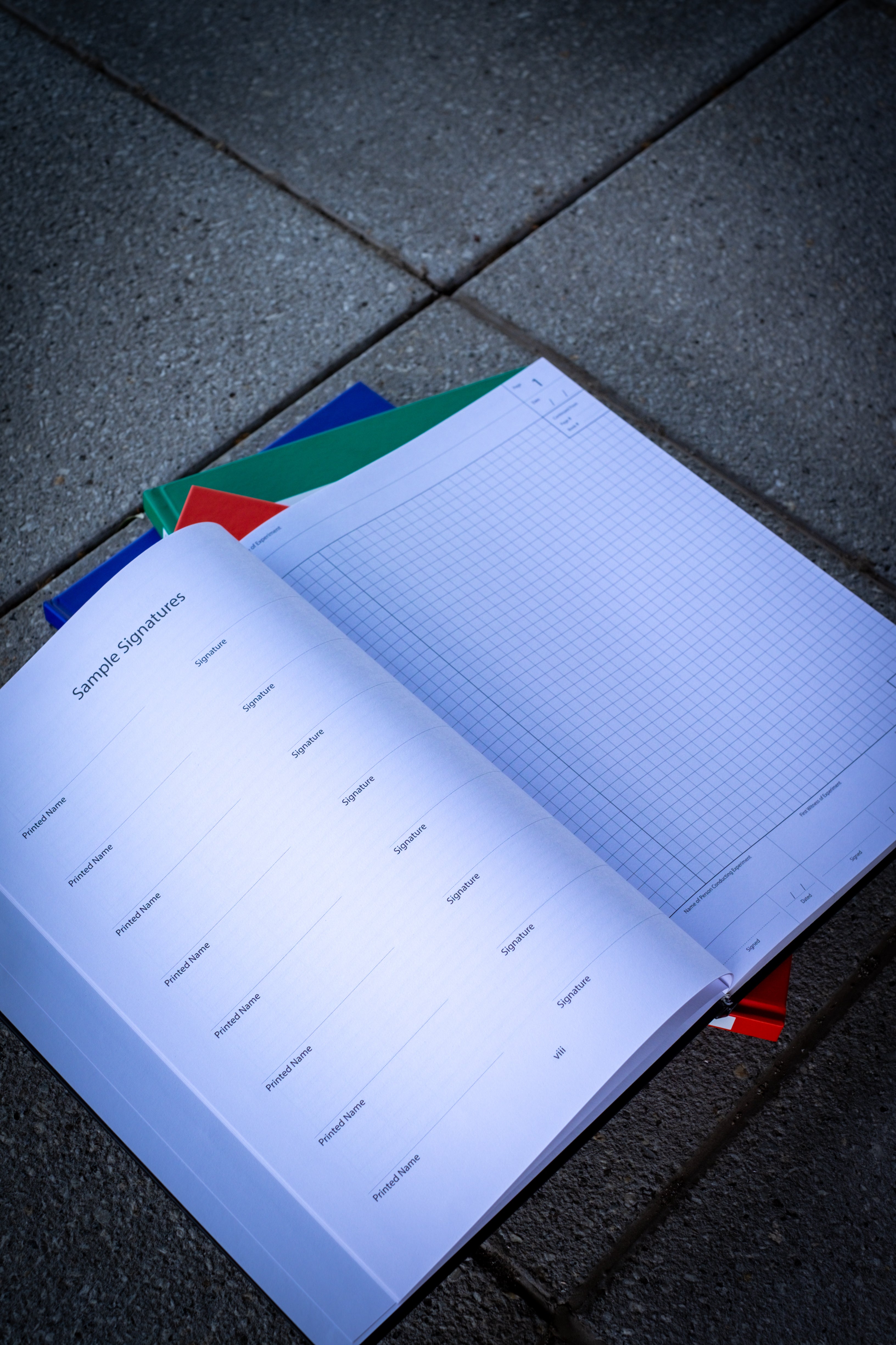 An opened black Laboratory Notebook placed on top of three closed notebooks (red, blue, and green) stacked underneath. The black notebook displays a grid page layout, emphasising the functional design for organising notes. The notebooks are arranged neatly on a black surface, highlighting the sleek and professional appearance of the notebook collection. 
