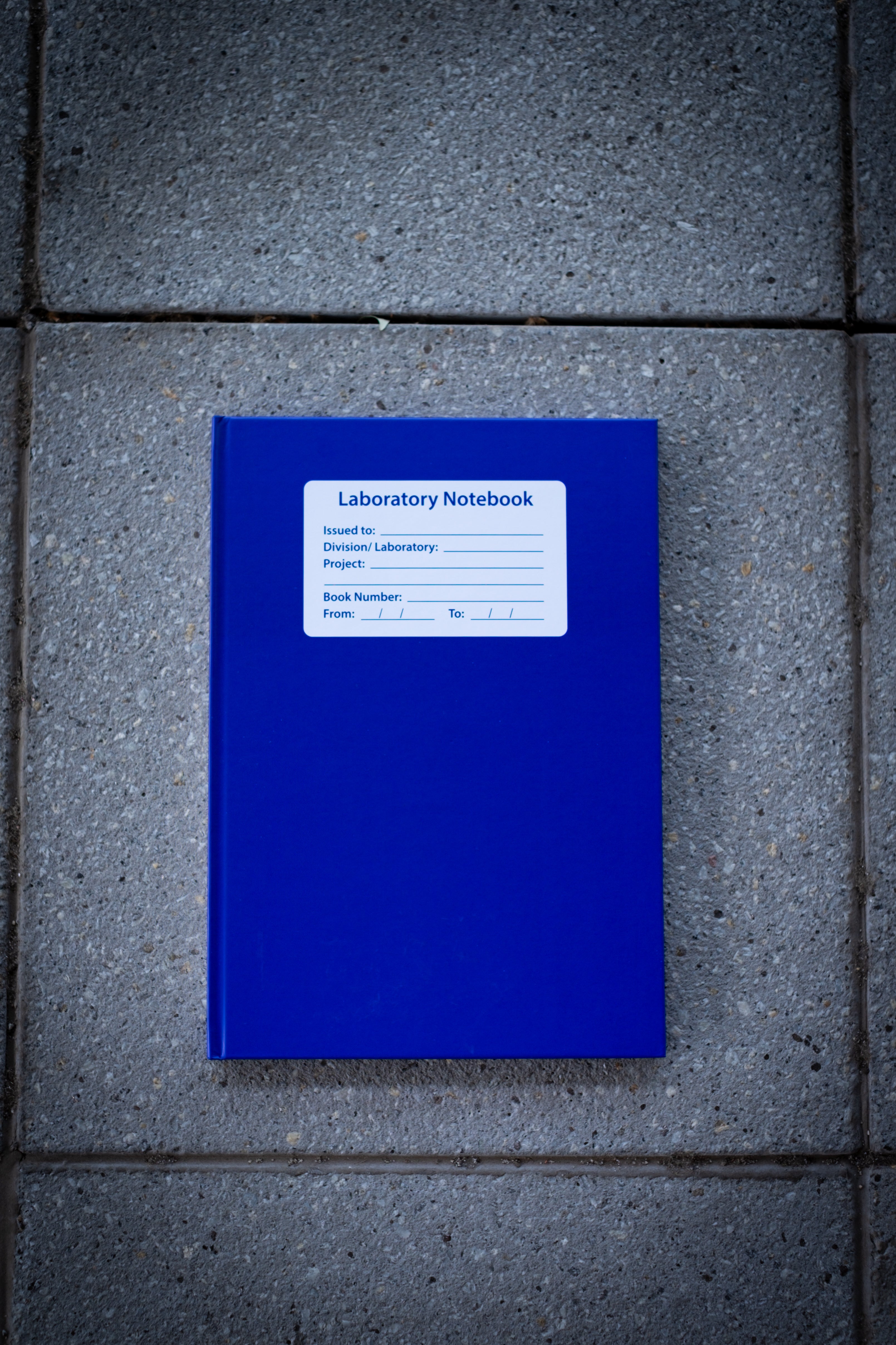 Laboratory Notebook in blue variant shown closed on a light surface, featuring a deep matte blue cover with rounded corners and a blank white rectangular label centred on the front — a durable, academic-use notebook available at RMIT Store.