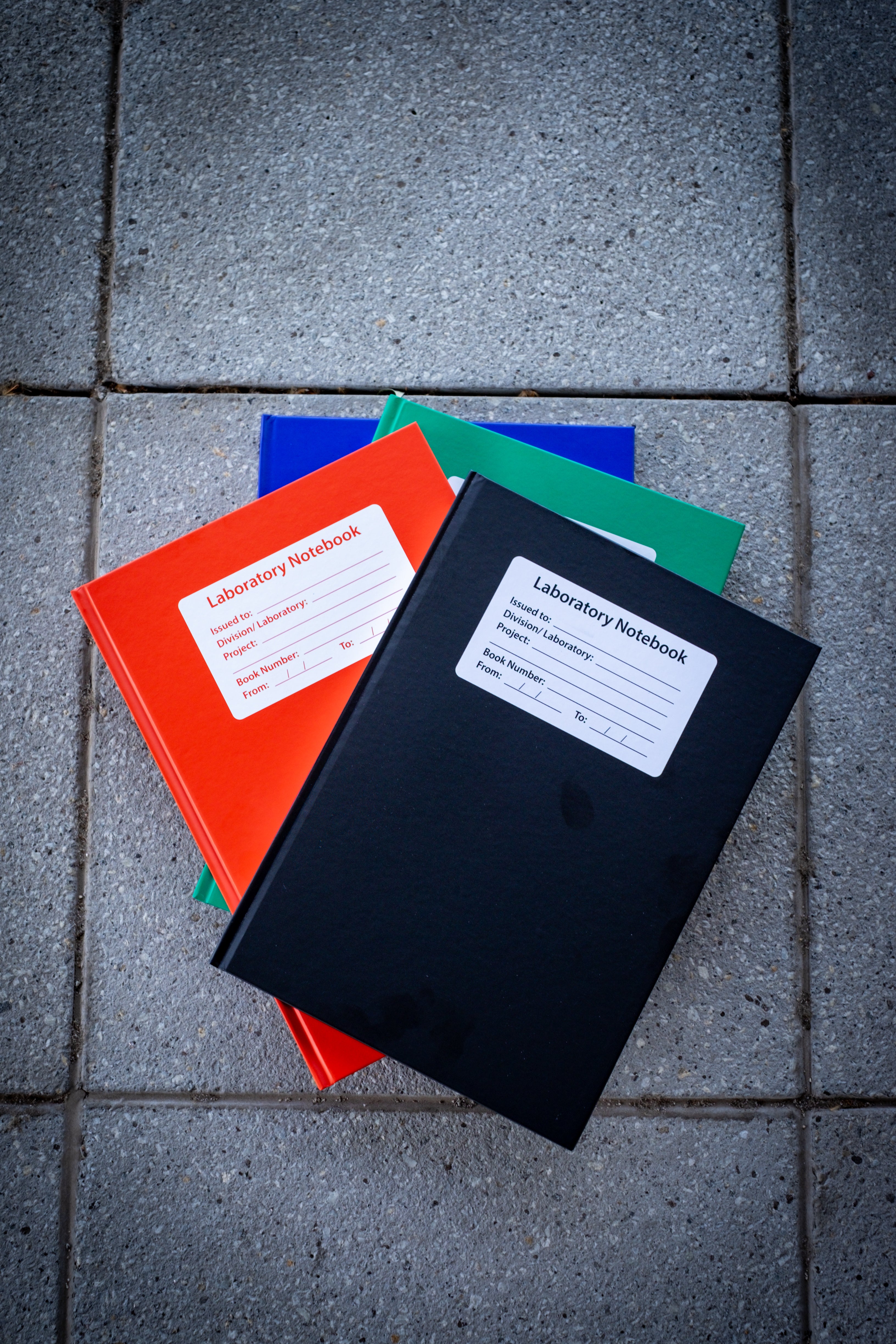 A collection of four closed Laboratory Notebooks stacked on top of each other, showcasing the different colour variants: black, red, blue, and green. Each notebook features a matte cover with a blank white rectangular label centred on the front for personalisation. Available at RMIT Store.