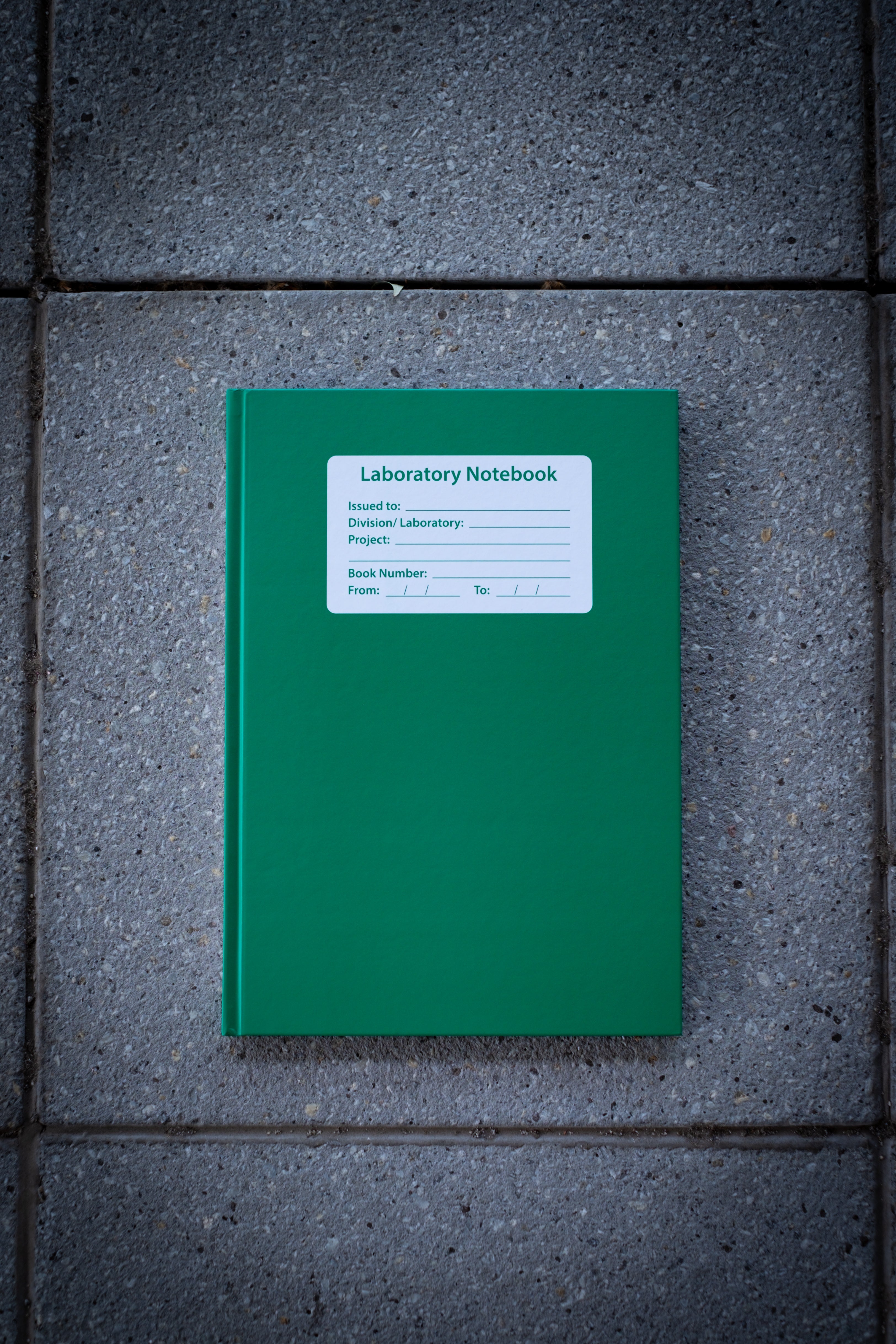 Laboratory Notebook in green variant displayed closed on a light surface, featuring a durable matte green cover with rounded corners and a white rectangular label at the centre for personalisation; image captured overhead in soft lighting to clearly show the front design — available at RMIT Store.