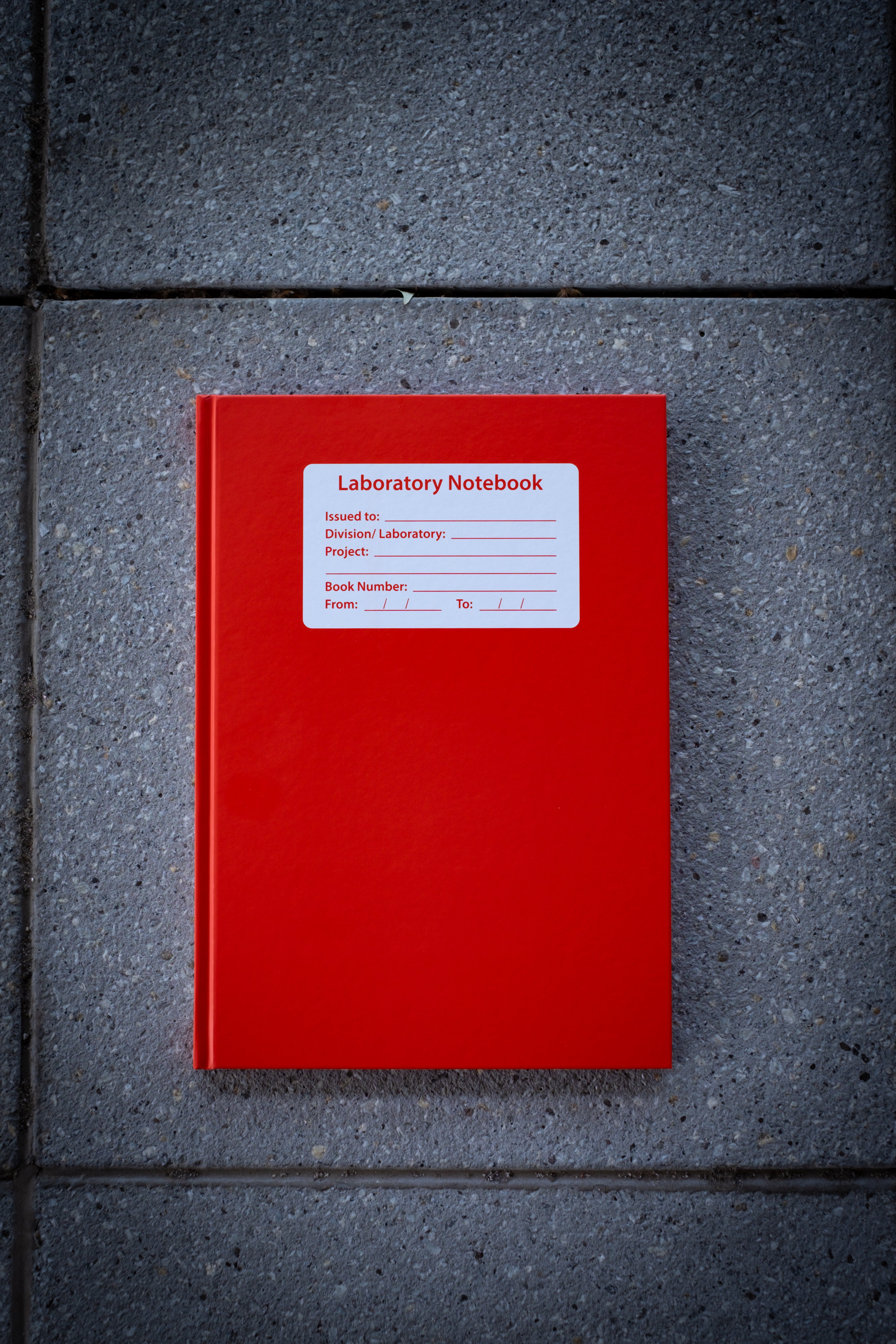 Laboratory Notebook in red variant shown closed on a light surface, featuring a bold matte red cover with rounded corners and a blank white rectangular label centred on the front, designed for research or academic use — available at RMIT Store.
