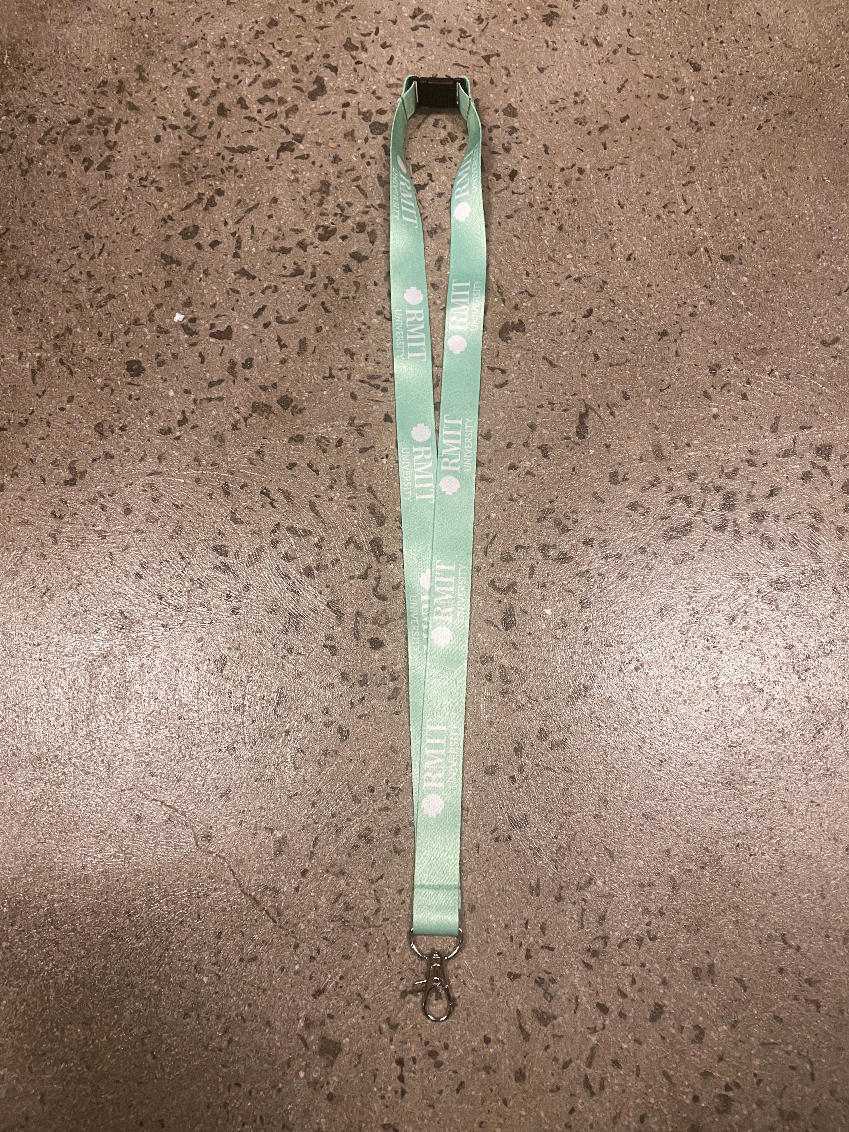 Green RMIT Store lanyard with the RMIT University logo, laid out flat on a textured surface. Designed with a silver clip for secure attachment, offering a practical accessory for identification.