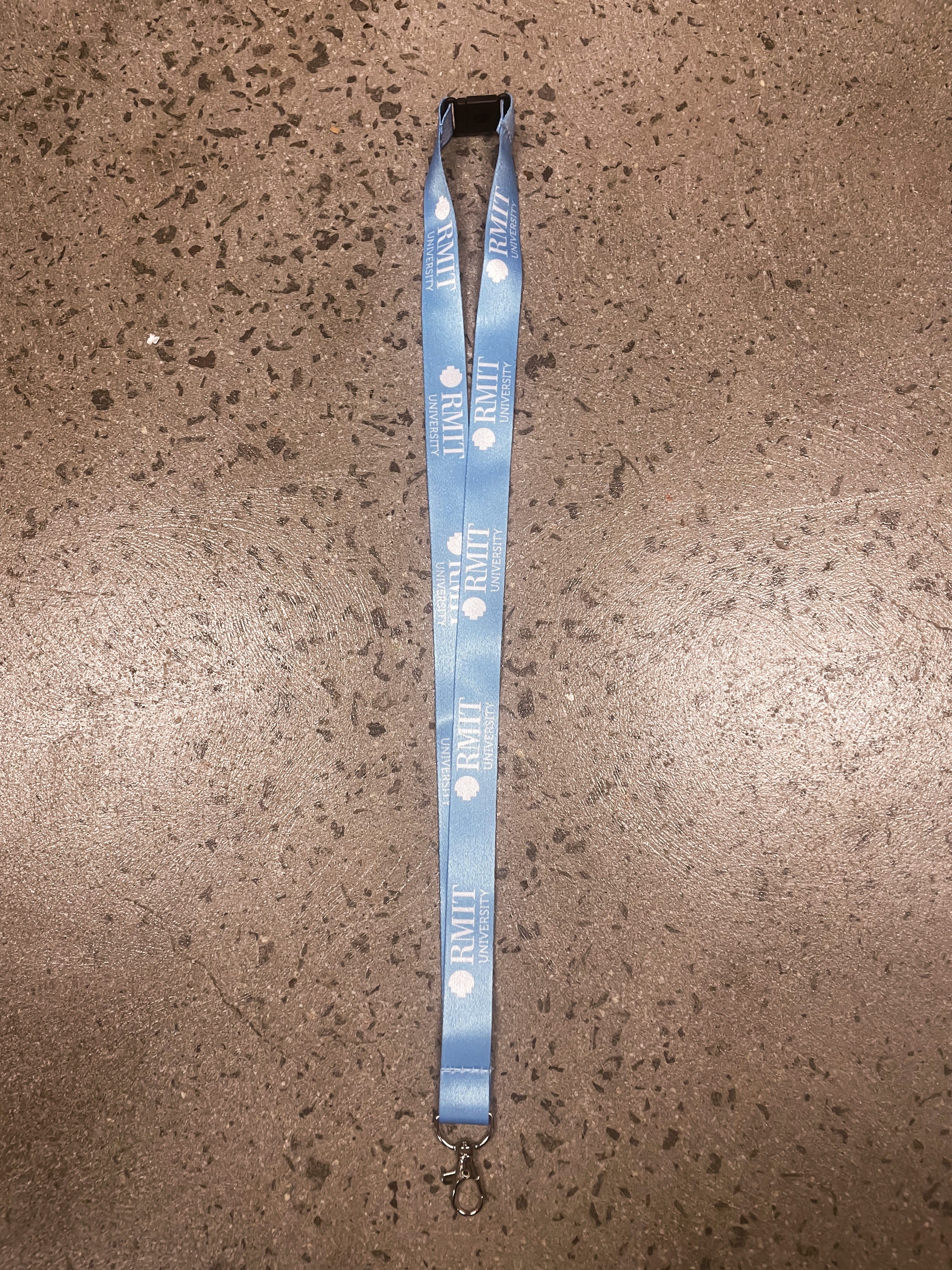 Light blue RMIT Store lanyard showcasing the RMIT University logo, equipped with a sturdy metal clip. Positioned flat against a light brown surface for clear visibility.