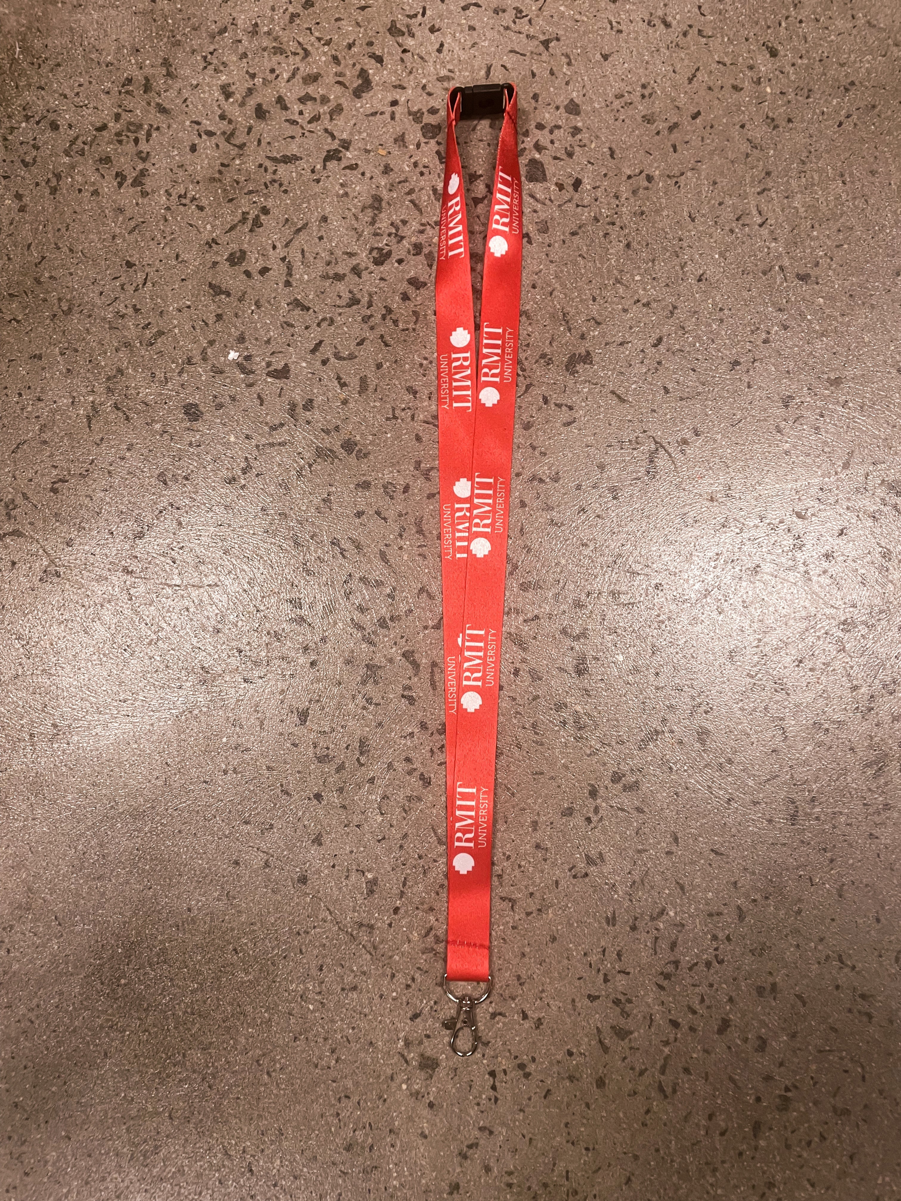 Red RMIT Store lanyard featuring the RMIT University logo, laid flat on a textured surface. Designed with a silver metal clip for secure attachment.