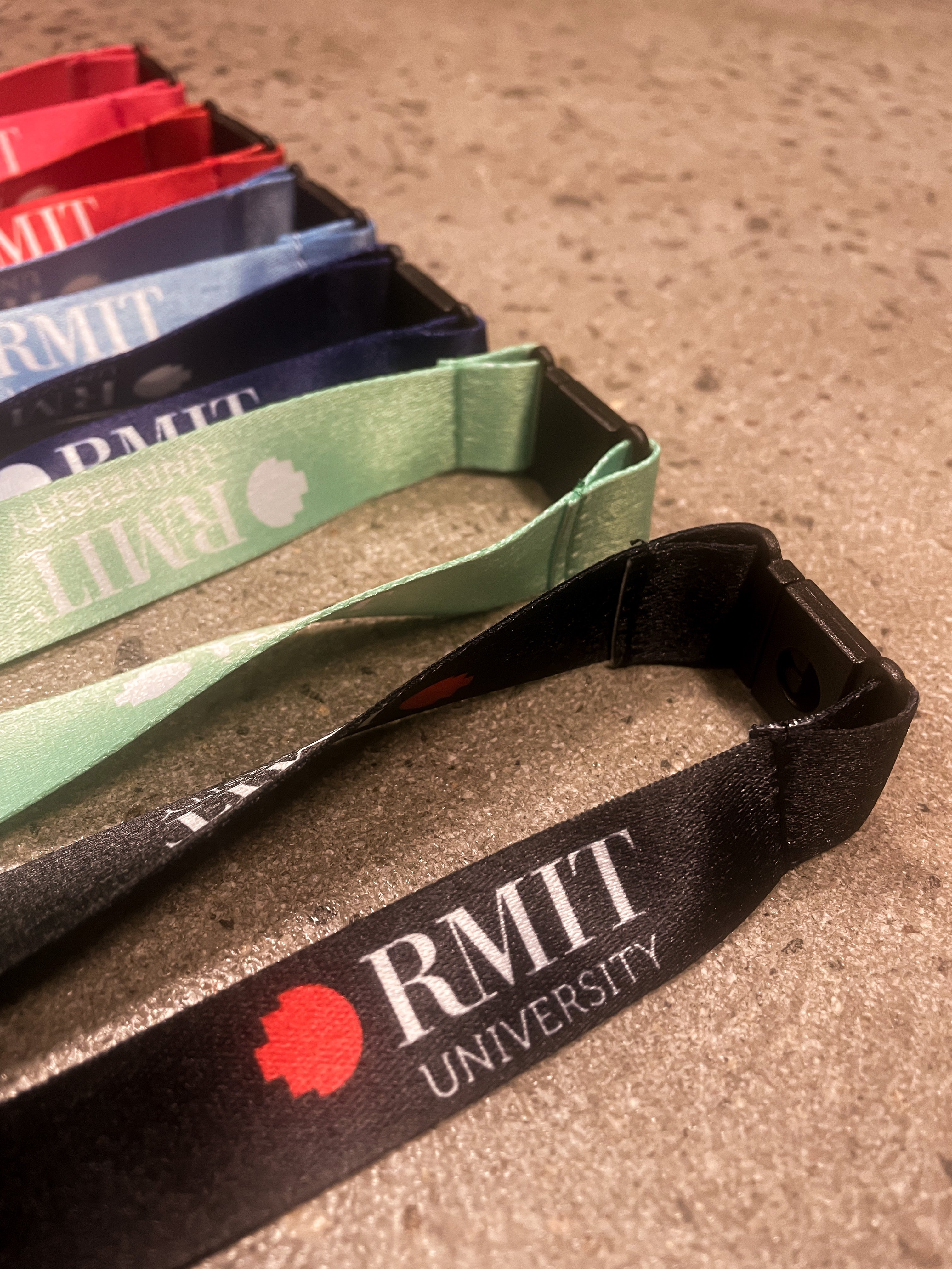 A close-up view of colourful RMIT Store lanyards displayed on a textured surface. The lanyards feature the RMIT University logo in varying shades: red, blue, green, and black, representing stylish and practical accessory options.