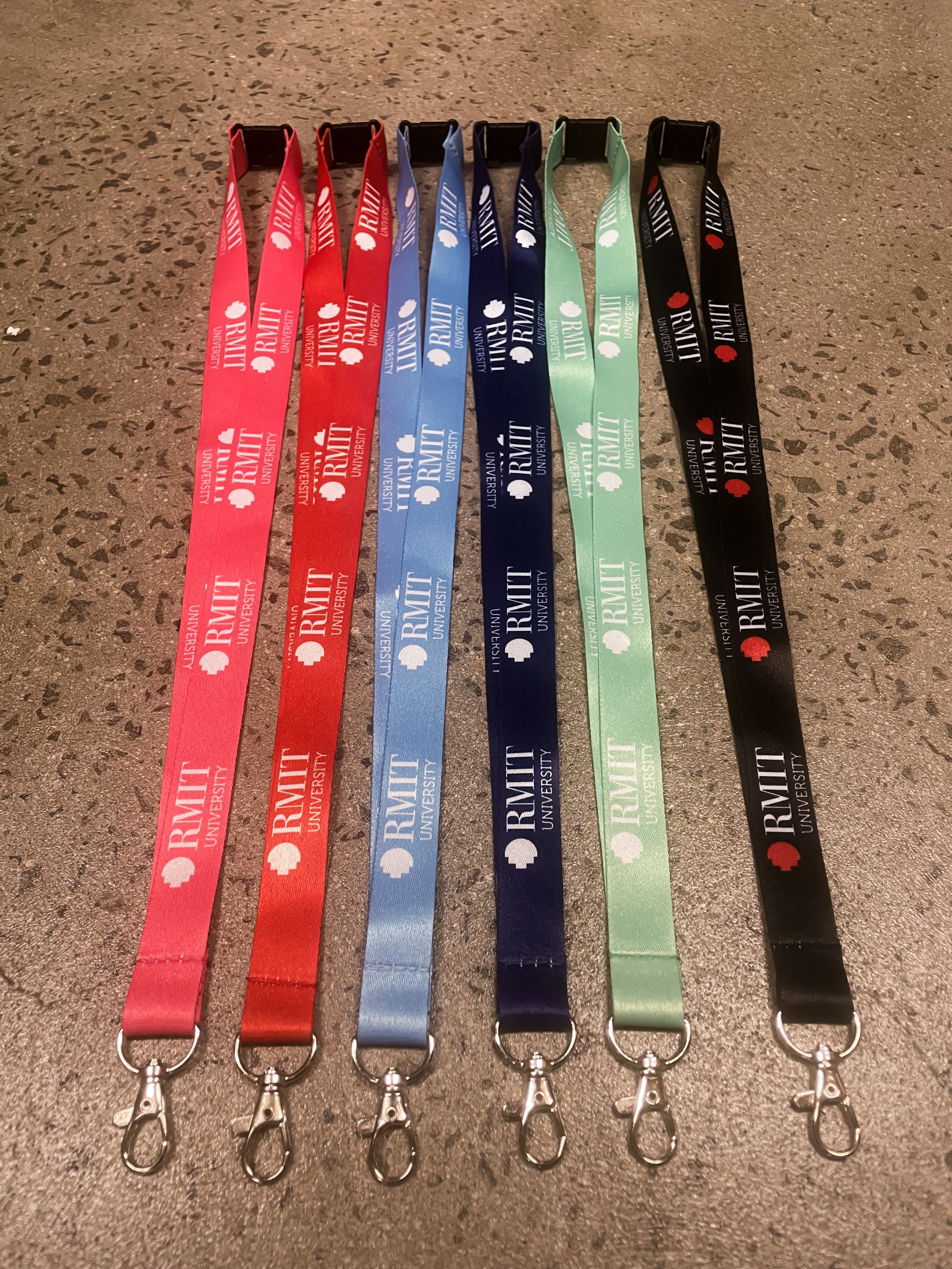 Six RMIT Store lanyards laid flat side by side in assorted colours: red, blue, green, and black. Each lanyard is adorned with the RMIT University logo and equipped with sturdy metal clips for secure attachment.