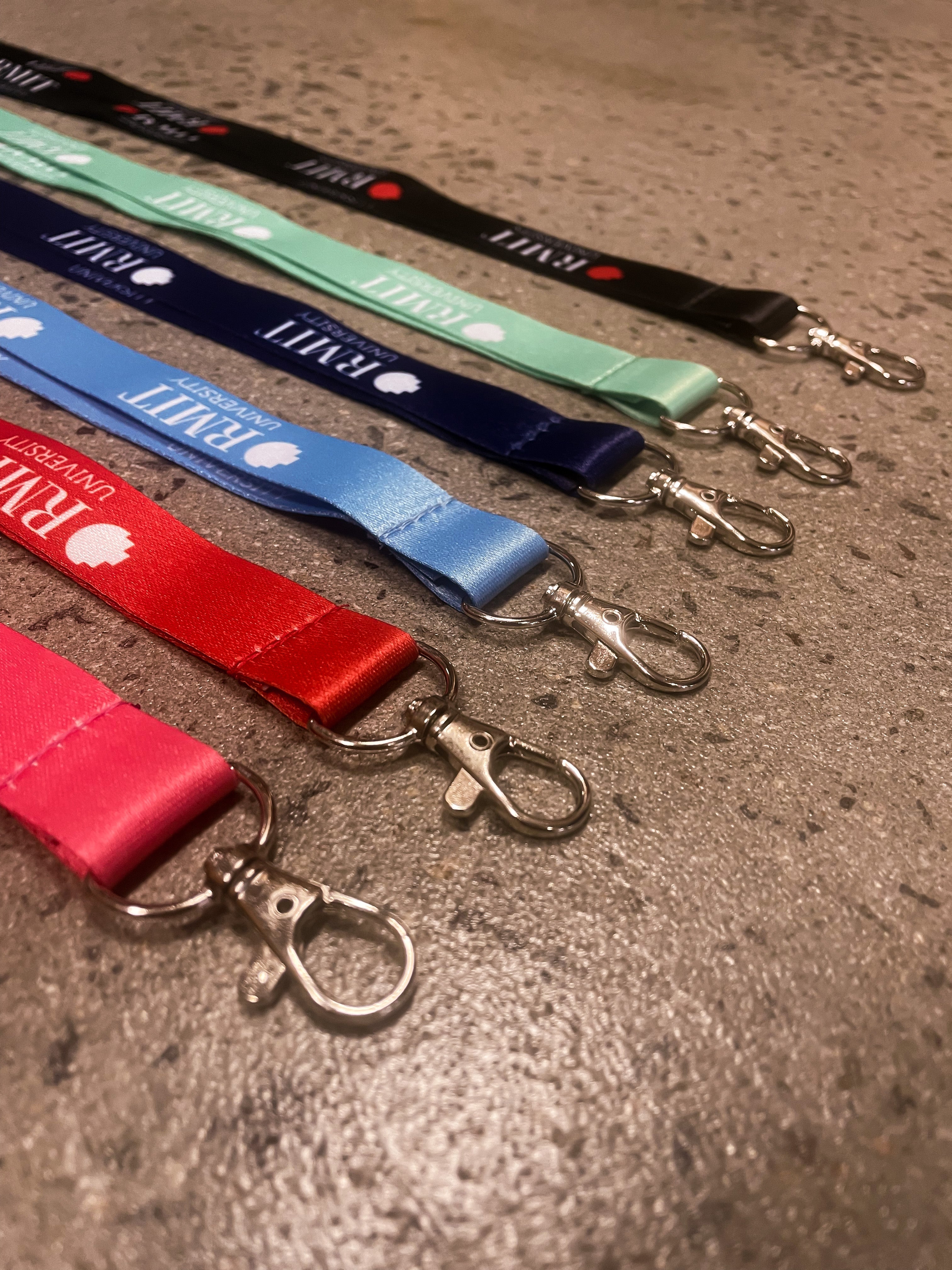 A series of RMIT Store lanyards in red, blue, green, and black, showcasing their silver metal clips on a smooth surface. Each lanyard displays the RMIT University logo, perfect for student and staff identification.