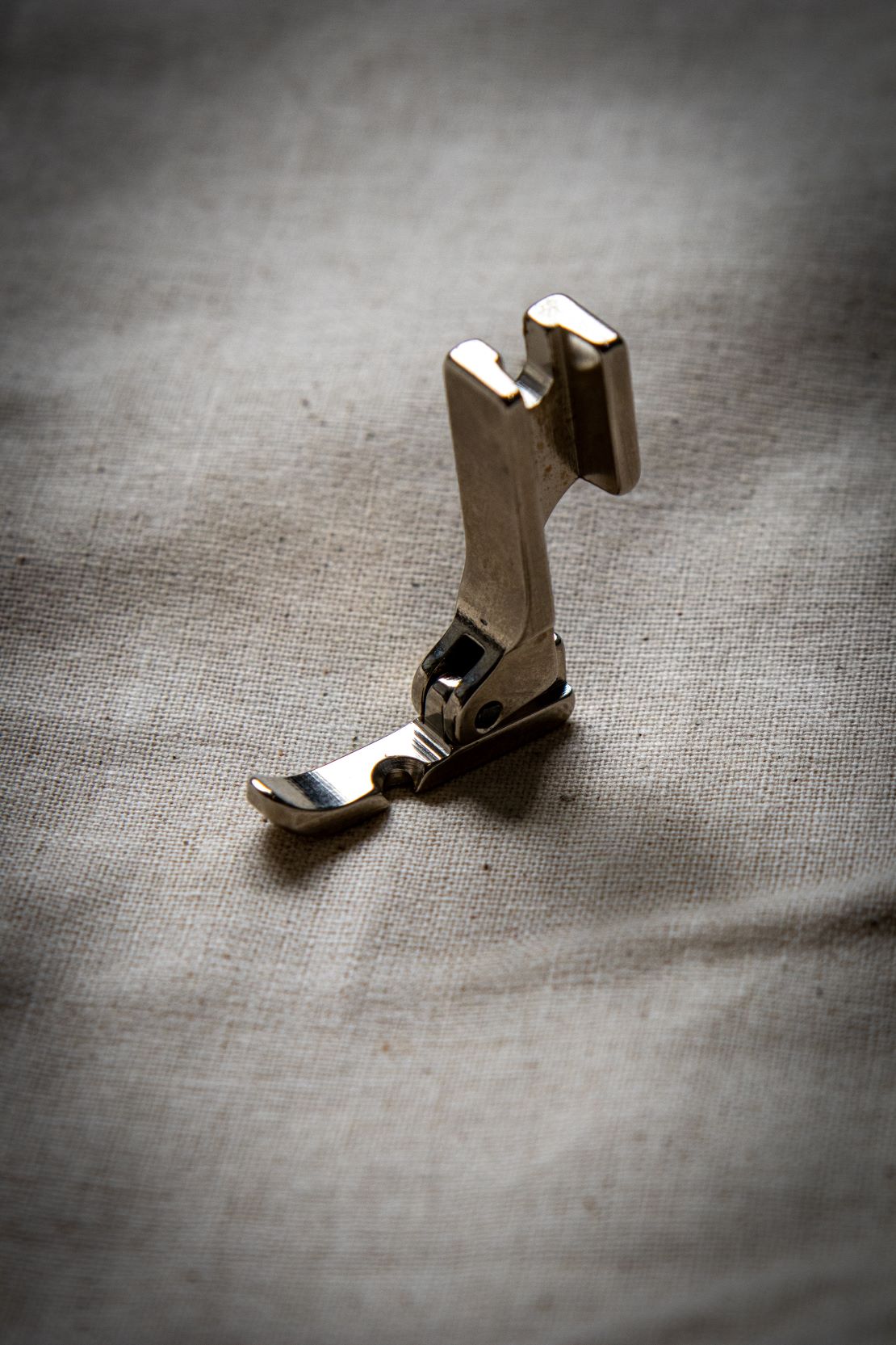 Close-up angled view of a silver left zipper foot sewing machine attachment resting on a light grey surface, clearly showing the left-positioned narrow channel and smooth curved base. Sold at the RMIT Store.