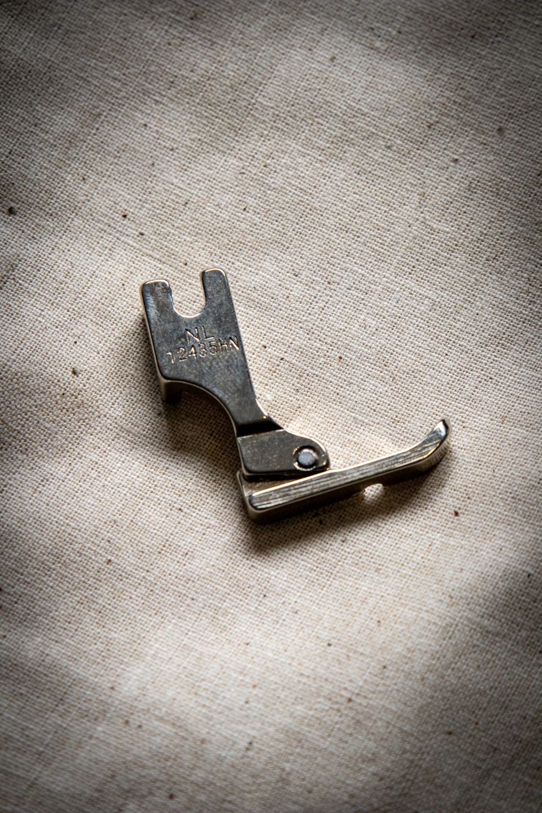 Side view of a silver left zipper foot sewing machine attachment on a pale grey background, showing the engraving "NL12435HN". Available at the RMIT Store.