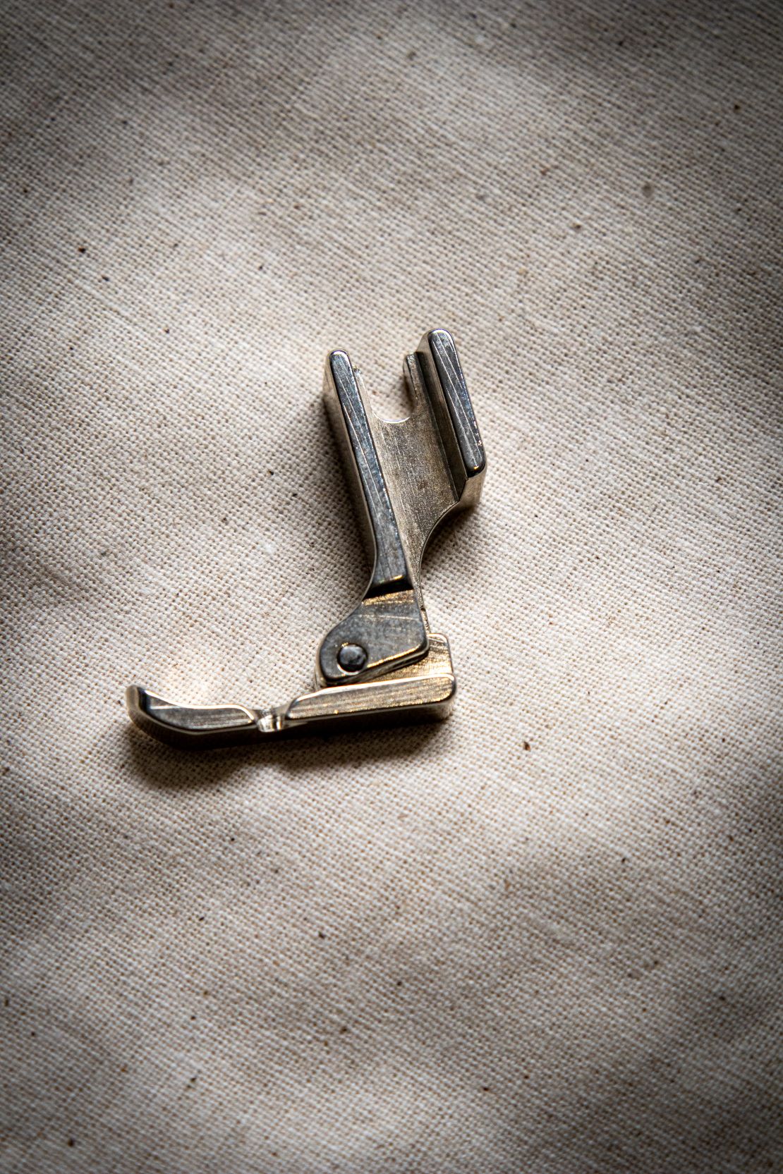 Top-down view of a silver left zipper foot sewing machine attachment placed on a smooth light grey surface, showing the left-side groove. Product stocked by the RMIT Store.