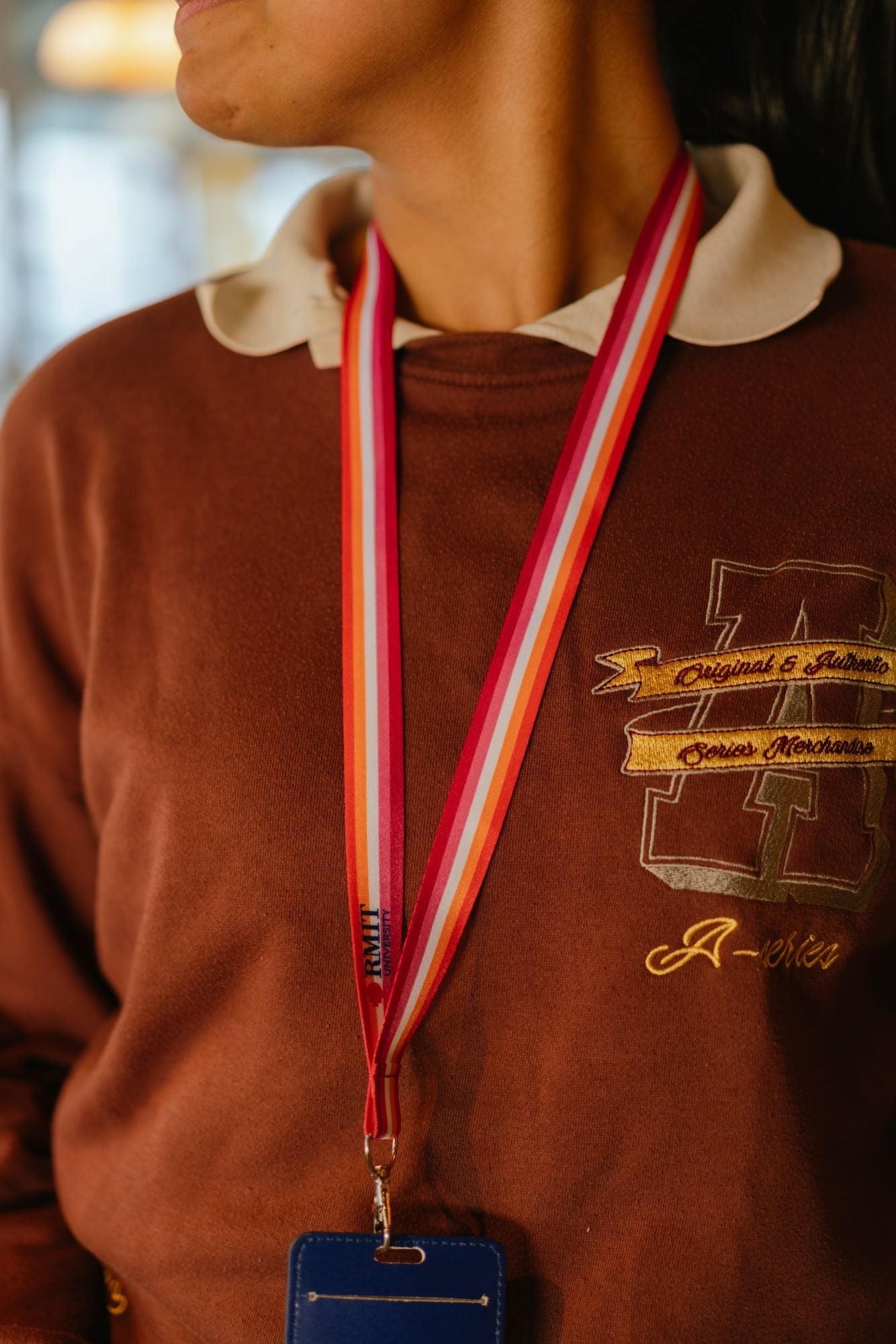 Close-up of a model wearing a Lesbian Pride lanyard from the RMIT Store, featuring horizontal stripes in dark orange, light orange, white, pink, and dark pink. The lanyard has a silver metal clip and a subtle engraved RMIT logo near the attachment point. The lanyard is attached to a card holder. The model wears an orange top.