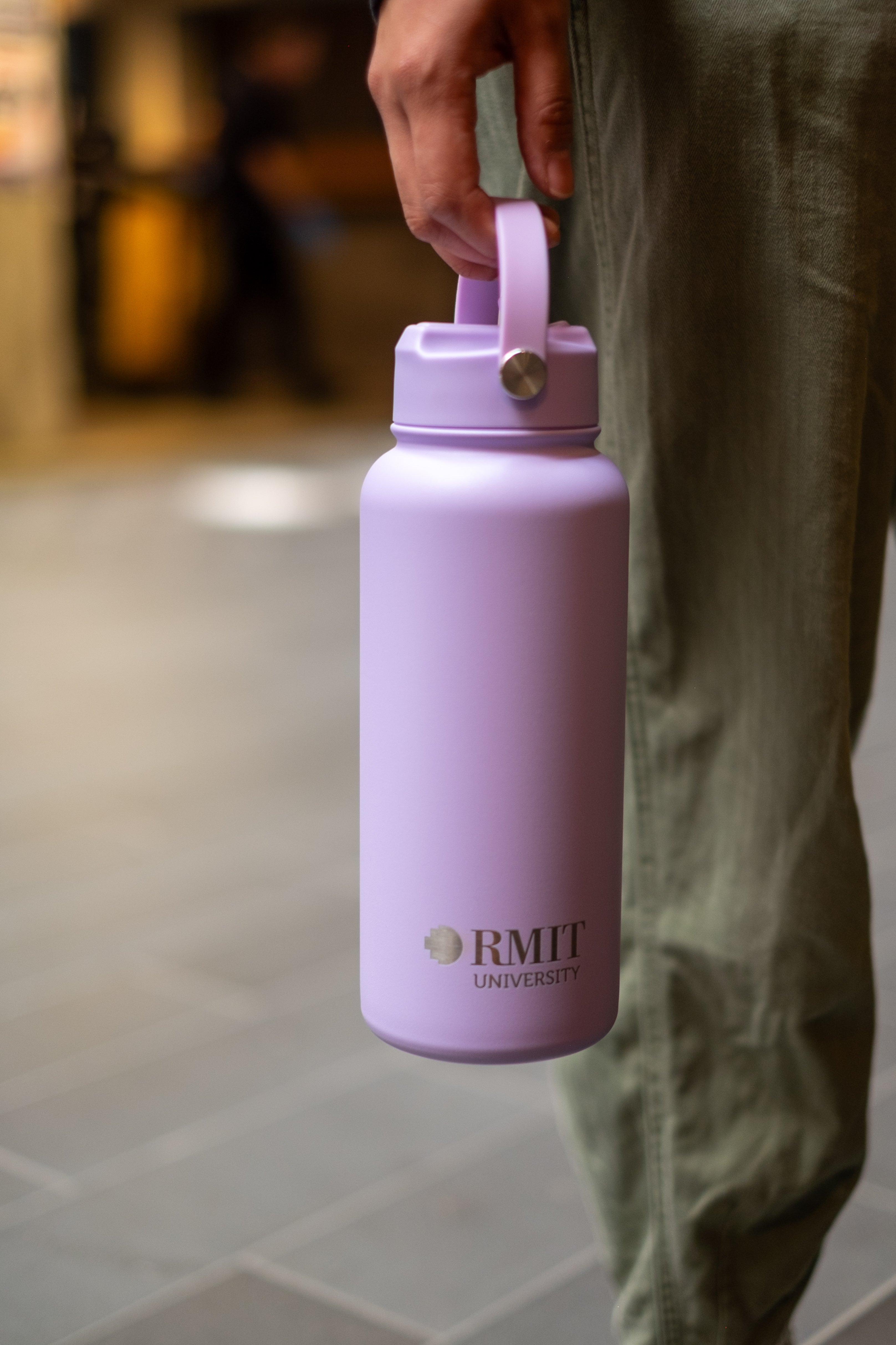 Person holding an RMIT Store Limited Edition Drink Bottle in Lavender, by the carry handle which is attached to the screw-on lid. The RMIT University logo is printed on the front in contrasting silver.