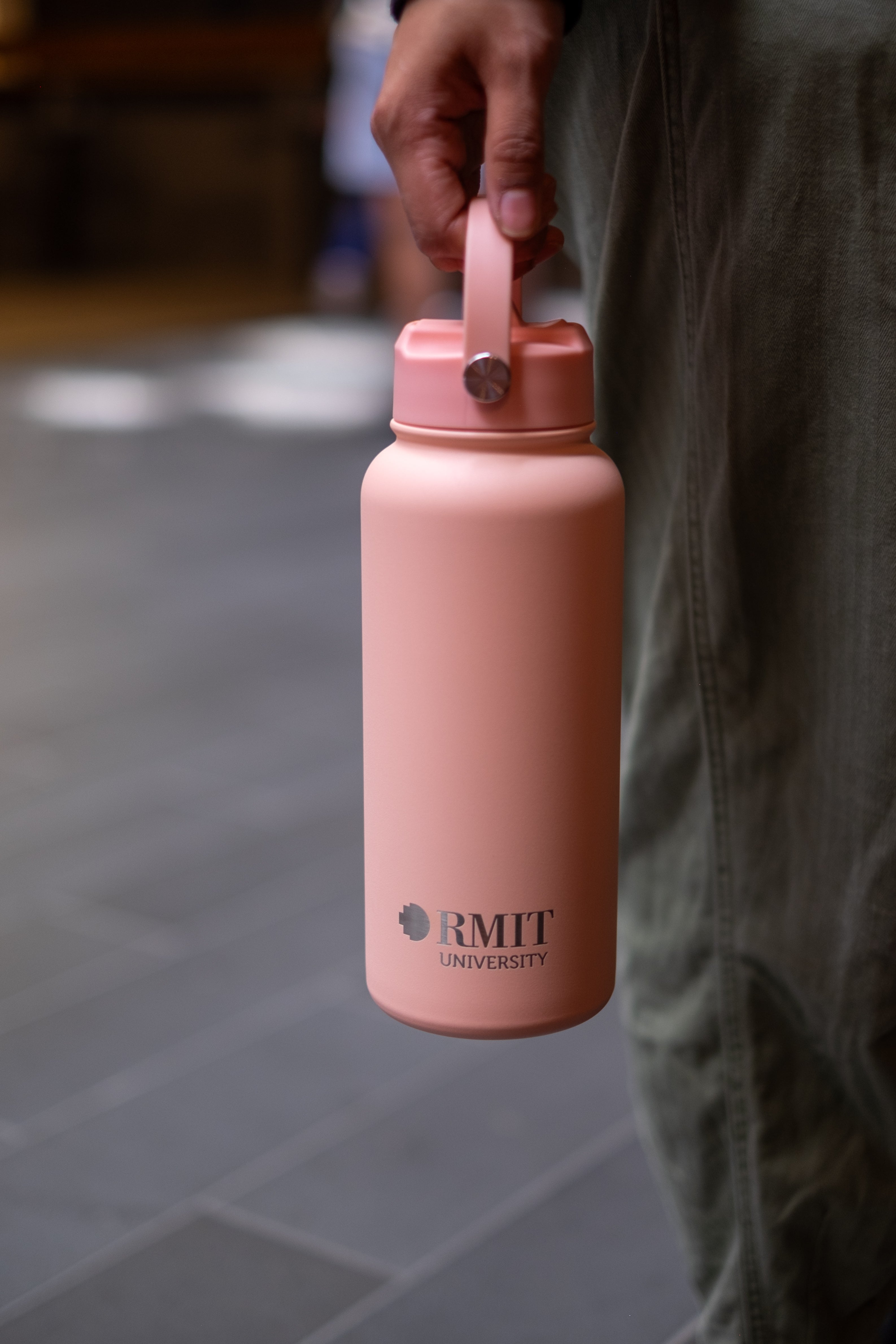 Person holding an RMIT Store Limited Edition Drink Bottle in Peach, by the carry handle which is attached to the screw-on lid. The RMIT University logo is printed on the front in contrasting silver.