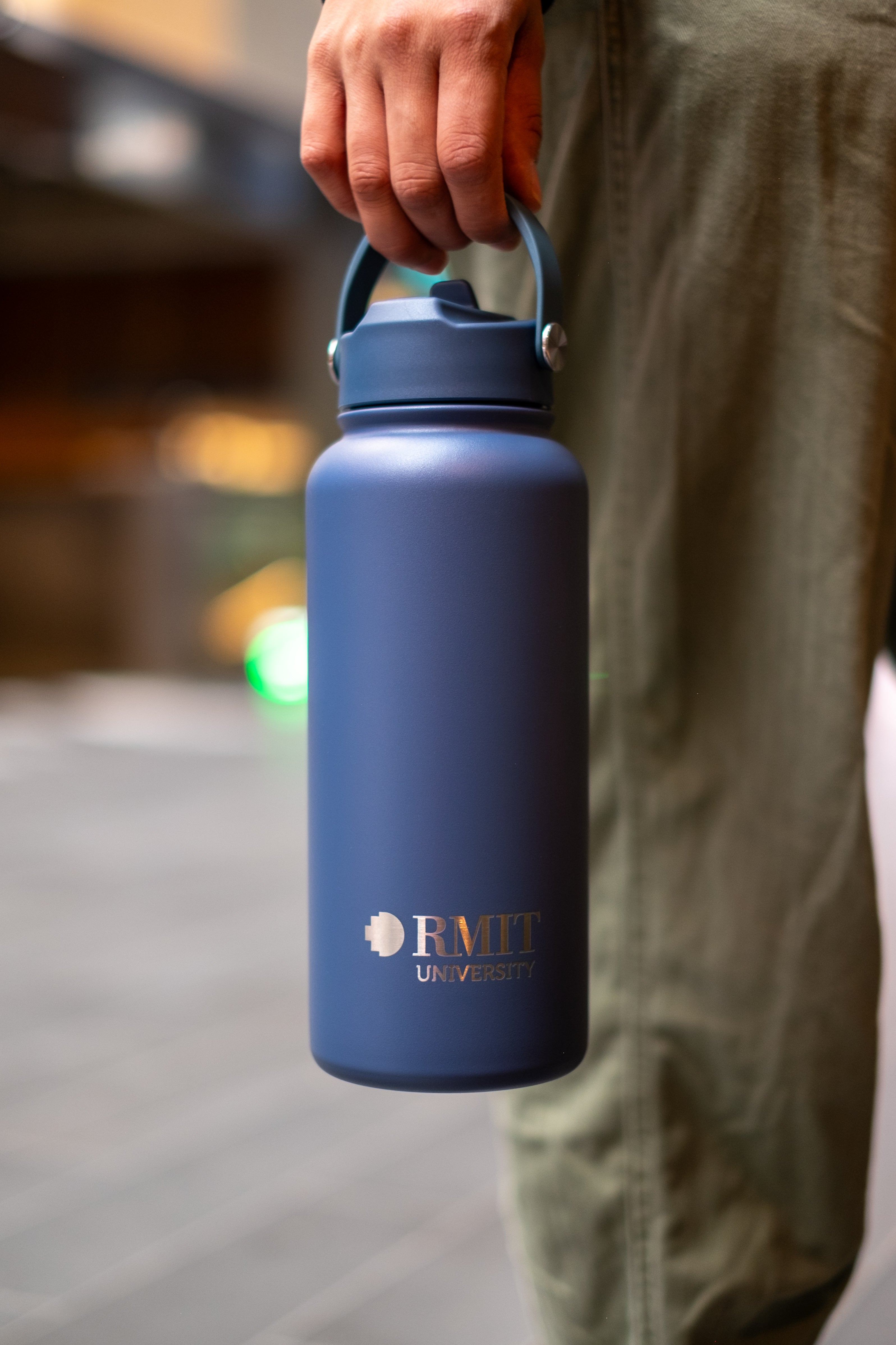 Person holding an RMIT Store Limited Edition Drink Bottle in Petrol Blue, by the carry handle which is attached to the screw-on lid. The RMIT University logo is printed on the front in contrasting silver.