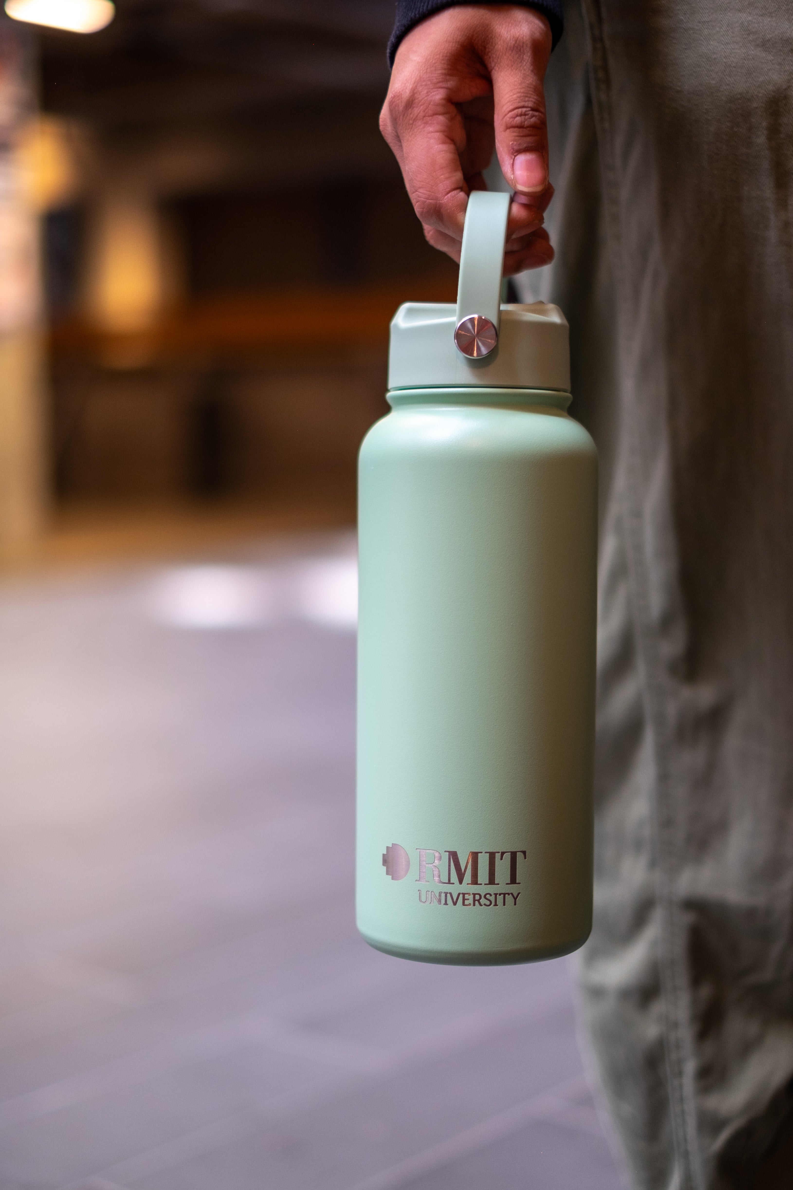Person holding an RMIT Store Limited Edition Drink Bottle in Sage, by the carry handle which is attached to the screw-on lid. The RMIT University logo is printed on the front in contrasting silver.