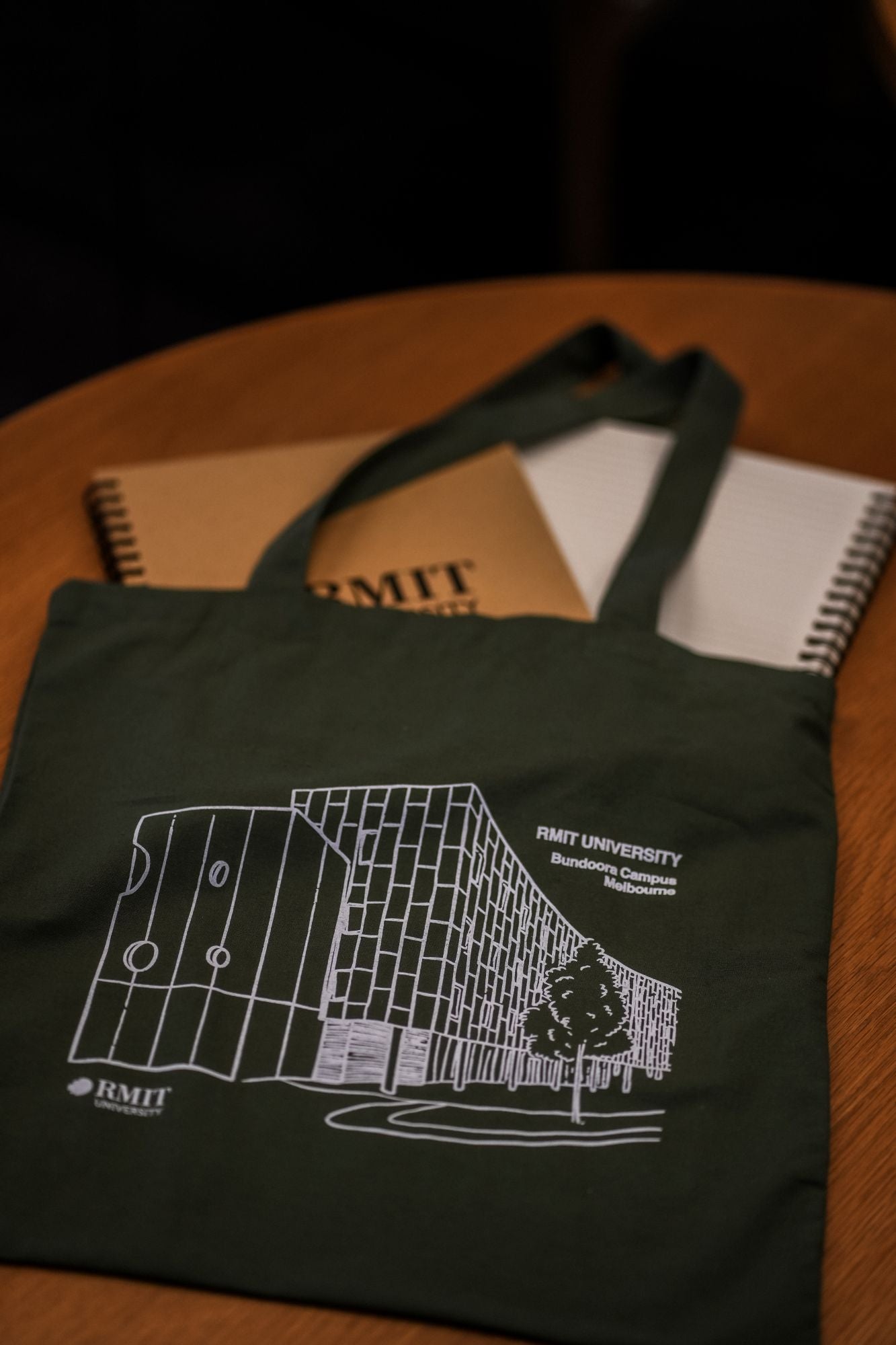 Olive Limited Edition Campus Tote with RMIT University Bundoora Campus design, laid flat on a light brown wooden table next to two RMIT-branded notebooks. The tote features a white line-art print of a contemporary campus building with grid-style windows and curved facade, along with a single tree illustration. Two printed logos are visible: “RMIT UNIVERSITY Bundoora Campus Melbourne” and the RMIT logo near the bottom left. Product available at the RMIT Store.