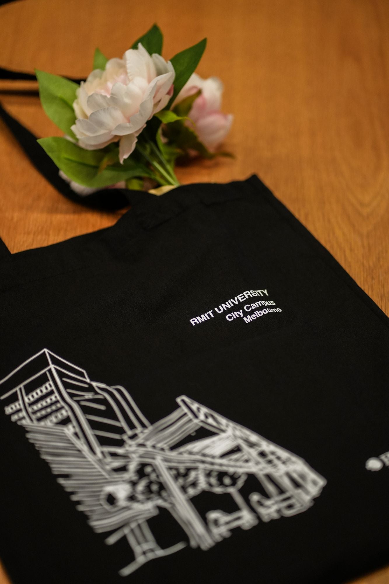 Black Limited Edition Campus Tote featuring RMIT University City Campus design, displayed on a light brown wooden surface with a bunch of pale pink artificial peonies partially tucked into the open top. The tote showcases a white line-art illustration of a Melbourne campus building with trees and architectural details, with two white printed logos: “RMIT UNIVERSITY City Campus Melbourne” near the top and the RMIT logo at the bottom right. Product available at the RMIT Store.