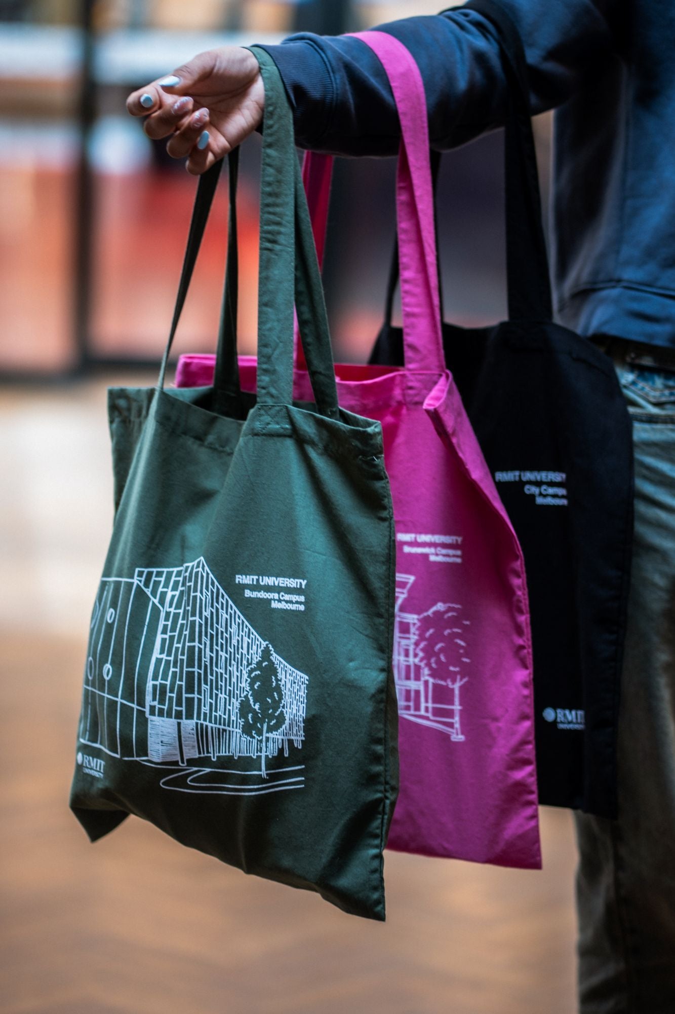 The Limited Edition Campus Tote collection featuring three tote bags in olive, pink, and black, each held by a model wearing a blue sweatshirt. The olive tote showcases an outline of RMIT University's Bundoora Campus, while the pink one displays the Brunswick Campus and the black tote highlights the City Campus. Each bag prominently features the RMIT University logo near the bottom corner of the prints. The model is indoors, grasping the totes by their handles, displaying the bags at hip level.