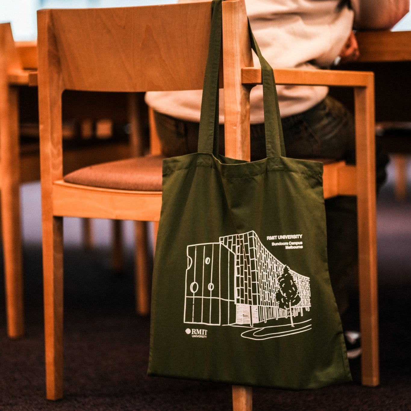 Dark olive green Limited Edition Campus Tote available at the RMIT Store, featuring a white line-art illustration of the RMIT University Bundoora Campus Melbourne building printed on the front with visible RMIT University logos and campus text branding. The cotton tote bag is modelled hanging from the back of a light timber chair in a warm indoor study or café setting, while a model wearing a light beige hoodie sits at a wooden table in the softly blurred background.