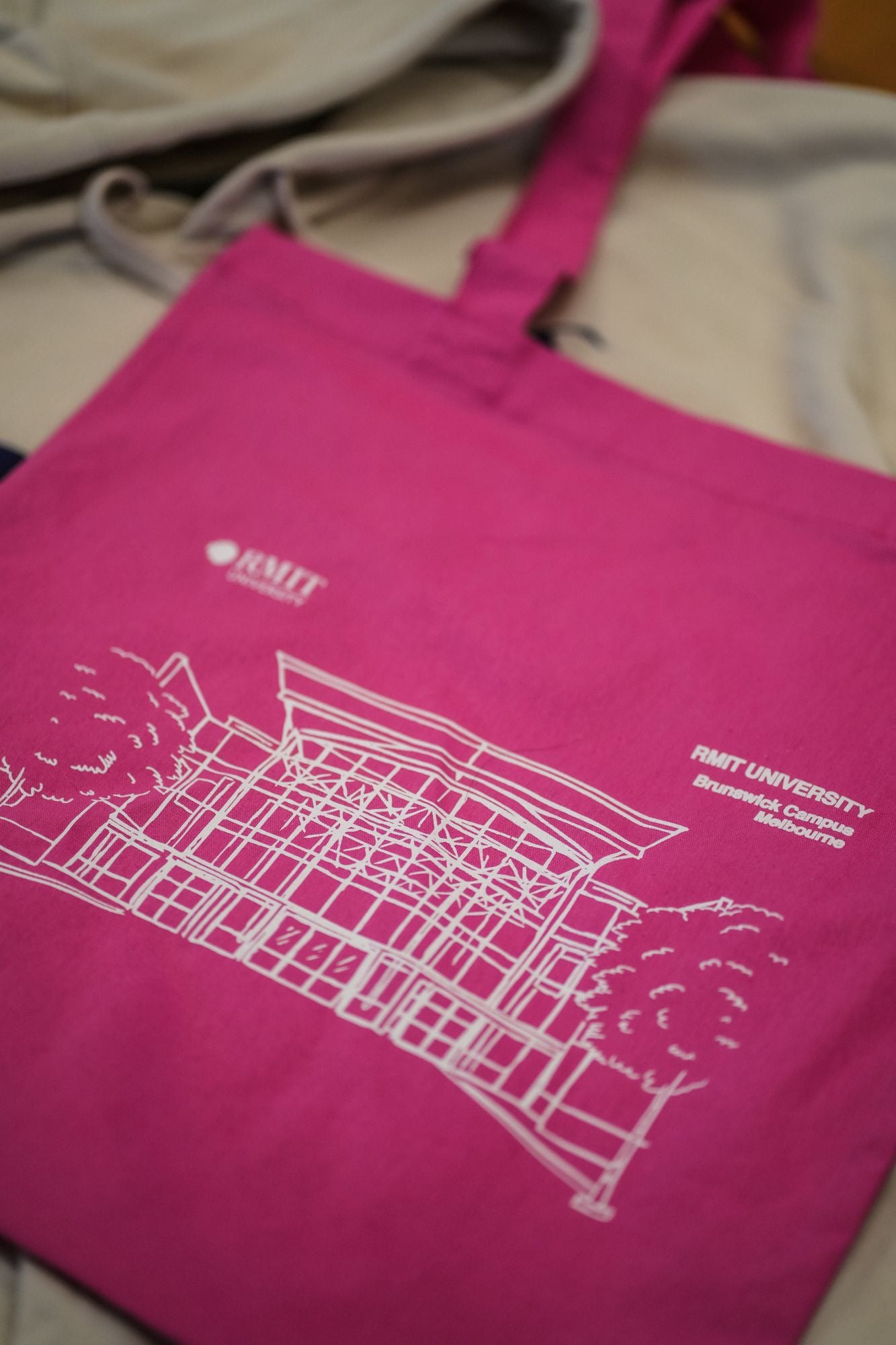 A vibrant pink Limited Edition Campus Tote showing the detailed outline of RMIT University's Brunswick Campus in white. The tote is laid on a soft beige hoodie, enhancing its colour contrast. The RMIT University logo and the "RMIT UNIVERSITY Brunswick Campus Melbourne" logo are alongside the Brunswick Campus print, showcasing architectural details including stylised buildings and trees. The image highlights the material and design intricacy of the tote from the RMIT Store.