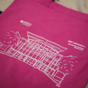 A vibrant pink Limited Edition Campus Tote showing the detailed outline of RMIT University's Brunswick Campus in white. The tote is laid on a soft beige hoodie, enhancing its colour contrast. The RMIT University logo and the 