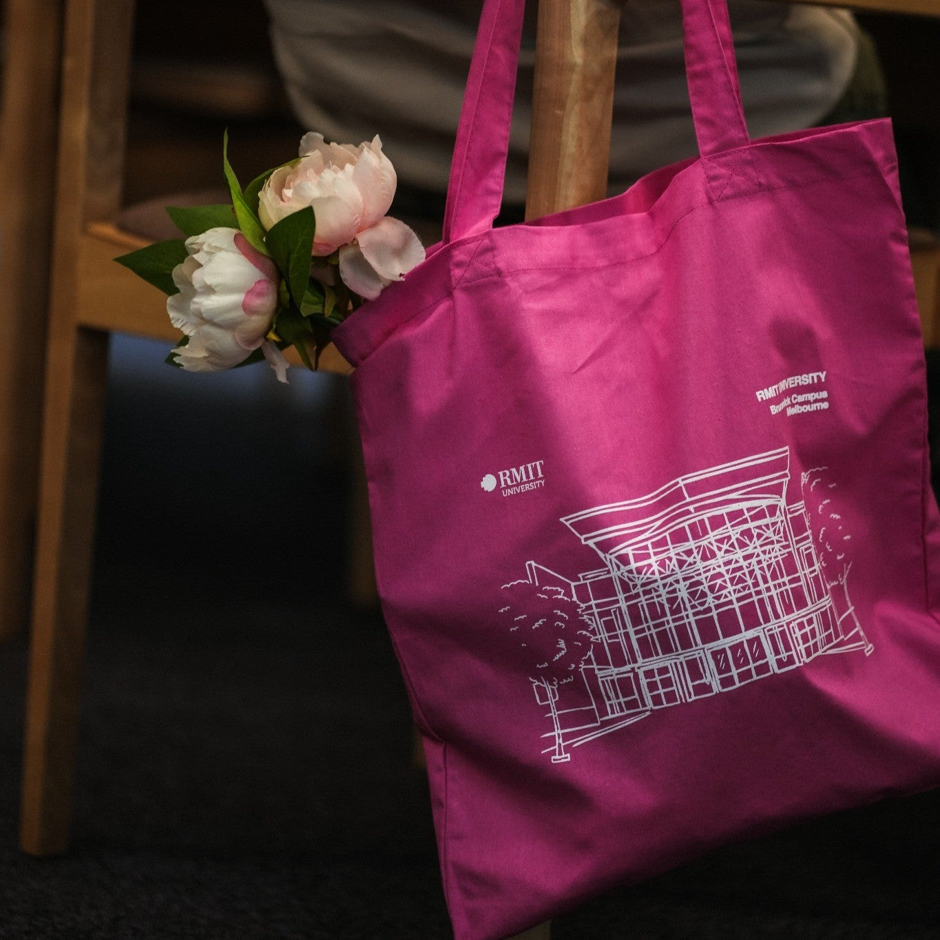 Bright pink Limited Edition Campus Tote available at the RMIT Store, featuring a white line-art illustration of the RMIT University Brunswick Campus Melbourne building on the front with visible RMIT University logos and campus text branding. The cotton tote bag is modelled hanging from the back of a wooden chair with soft pink flowers placed inside the bag, while a model wearing a grey hoodie sits at a table in a warm indoor café or campus environment with softly blurred background lighting.