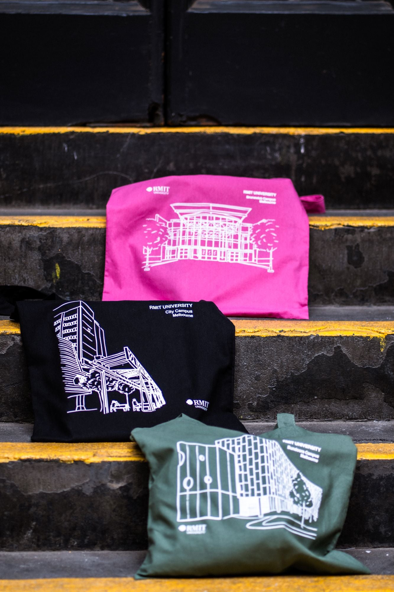 Three Limited Edition Campus Totes in pink, black, and olive, arranged on concrete steps. The pink tote features the Brunswick Campus design, the black tote highlights the City Campus, and the olive tote showcases the Bundoora Campus, all with the RMIT University logo. The totes are neatly displayed, illustrating their visibility and style against the rugged backdrop of urban steps, signifying a casual and accessible design from the RMIT Store.