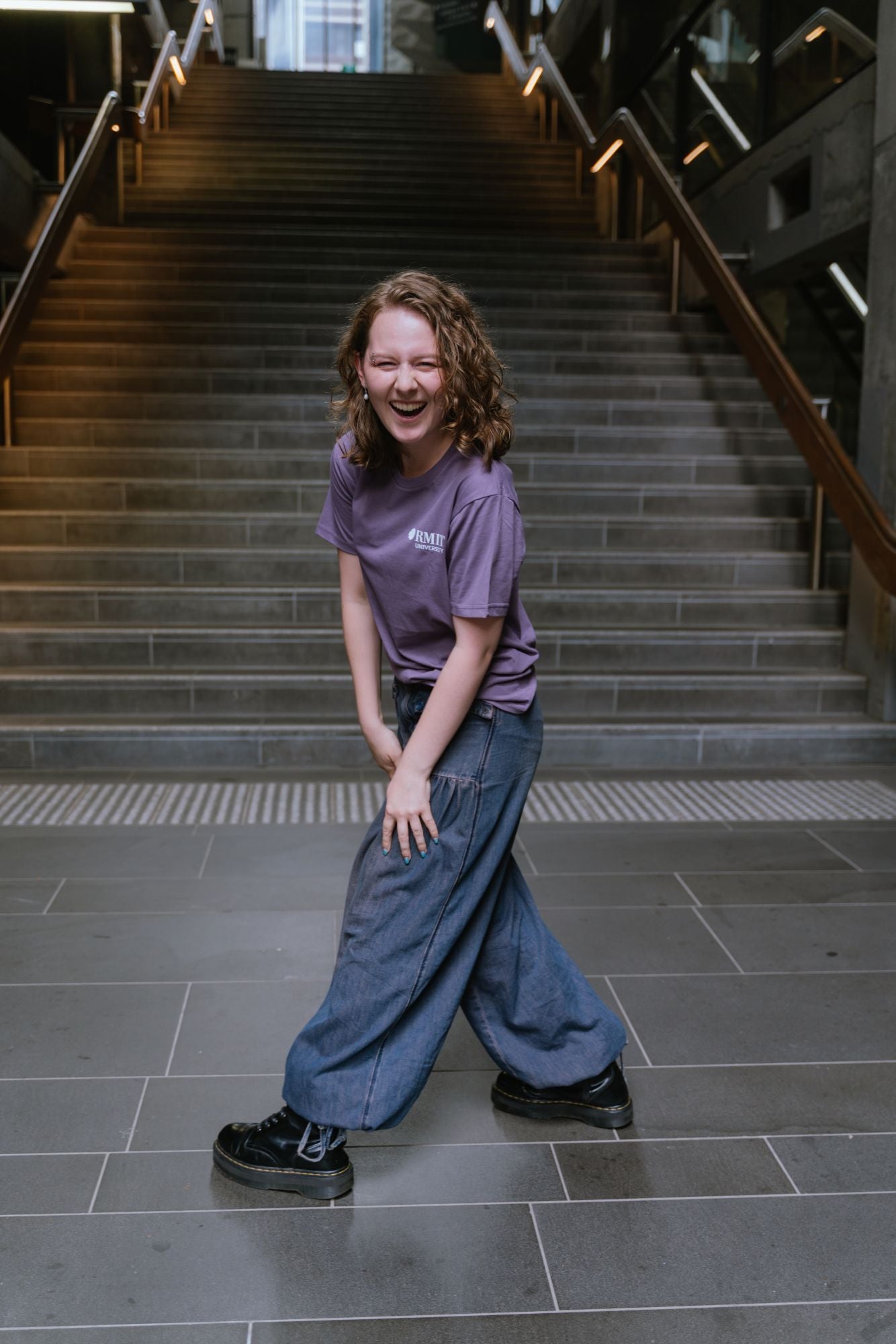 Model standing in indoor stairwell wearing Limited Edition Tees ’26 in mauve with small white RMIT University logo on the left chest, styled with dark denim wide-leg jeans and black boots, laughing while leaning slightly forward with playful pose, available at the RMIT Store.