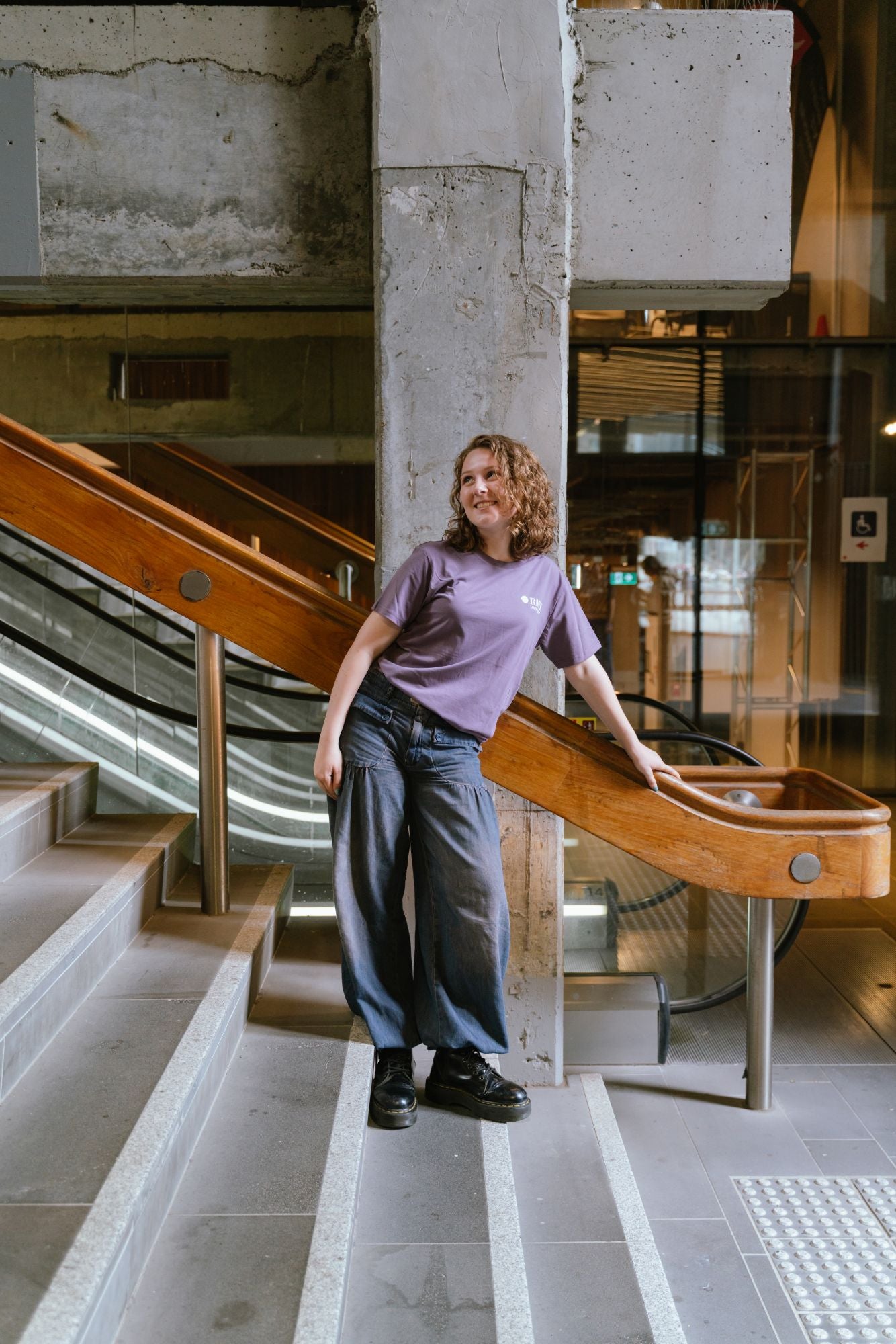 Model standing indoors on a staircase landing wearing Limited Edition Tees ’26 in mauve featuring a small white RMIT University logo printed on the left chest, paired with loose dark denim wide-leg jeans and black lace-up boots, leaning against a wooden handrail with industrial concrete and escalator background, available at the RMIT Store.